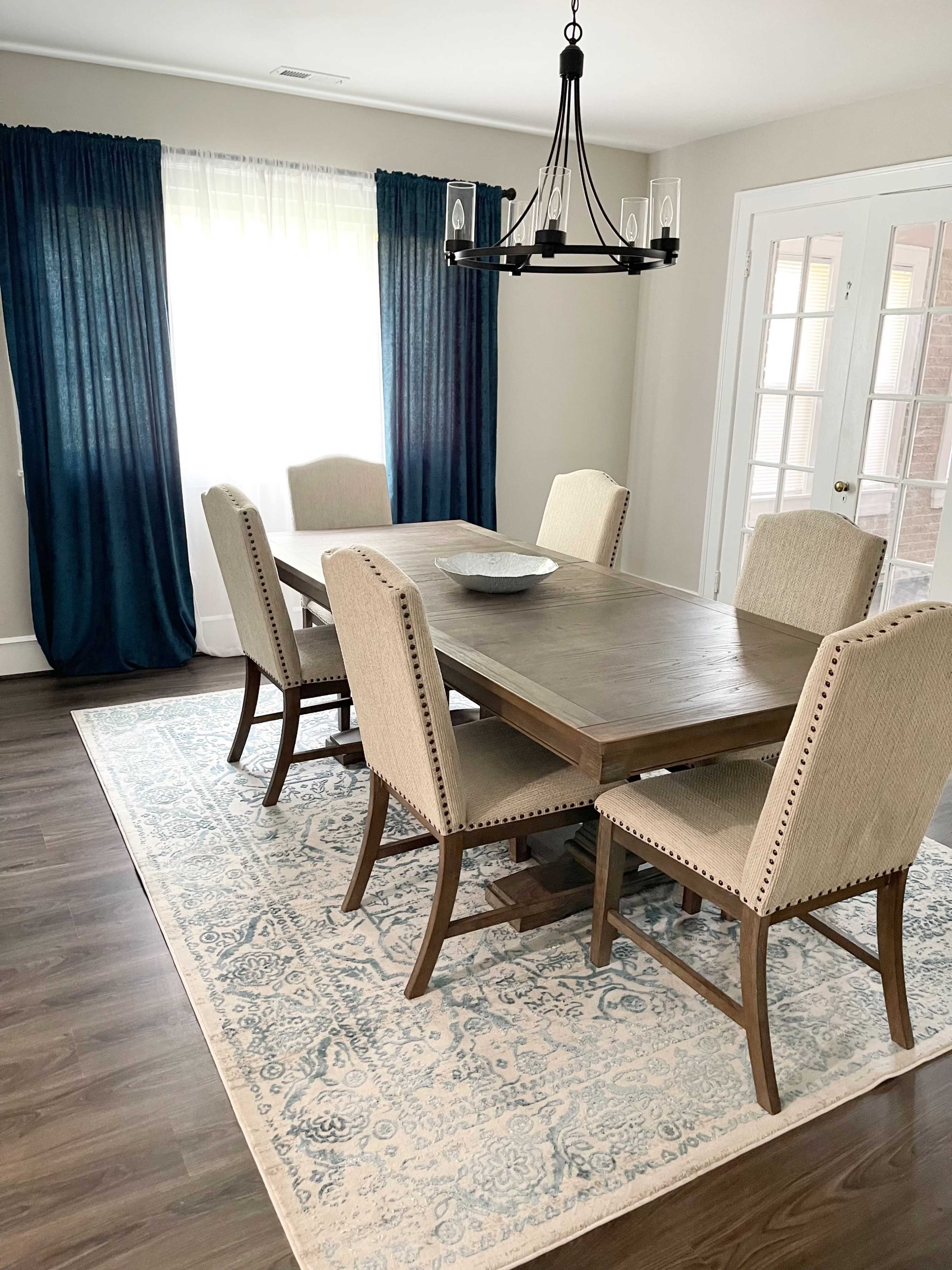 A dining room features a wooden table with six upholstered chairs, a large area rug, and a chandelier above, complemented by sheer curtains.