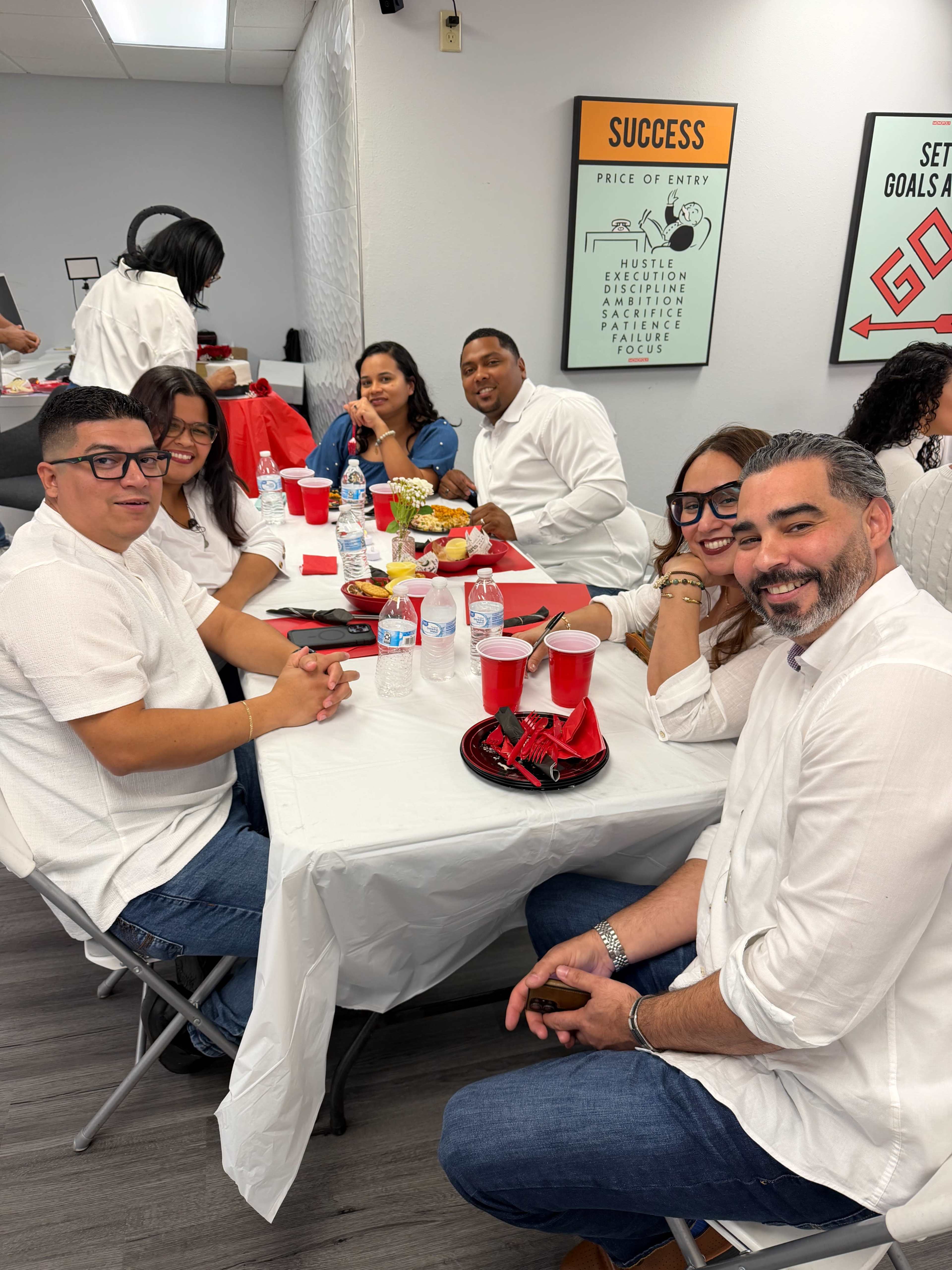 A group of six people sits at a table covered with a white tablecloth, enjoying food and drinks in a brightly lit room decorated with motivational posters.