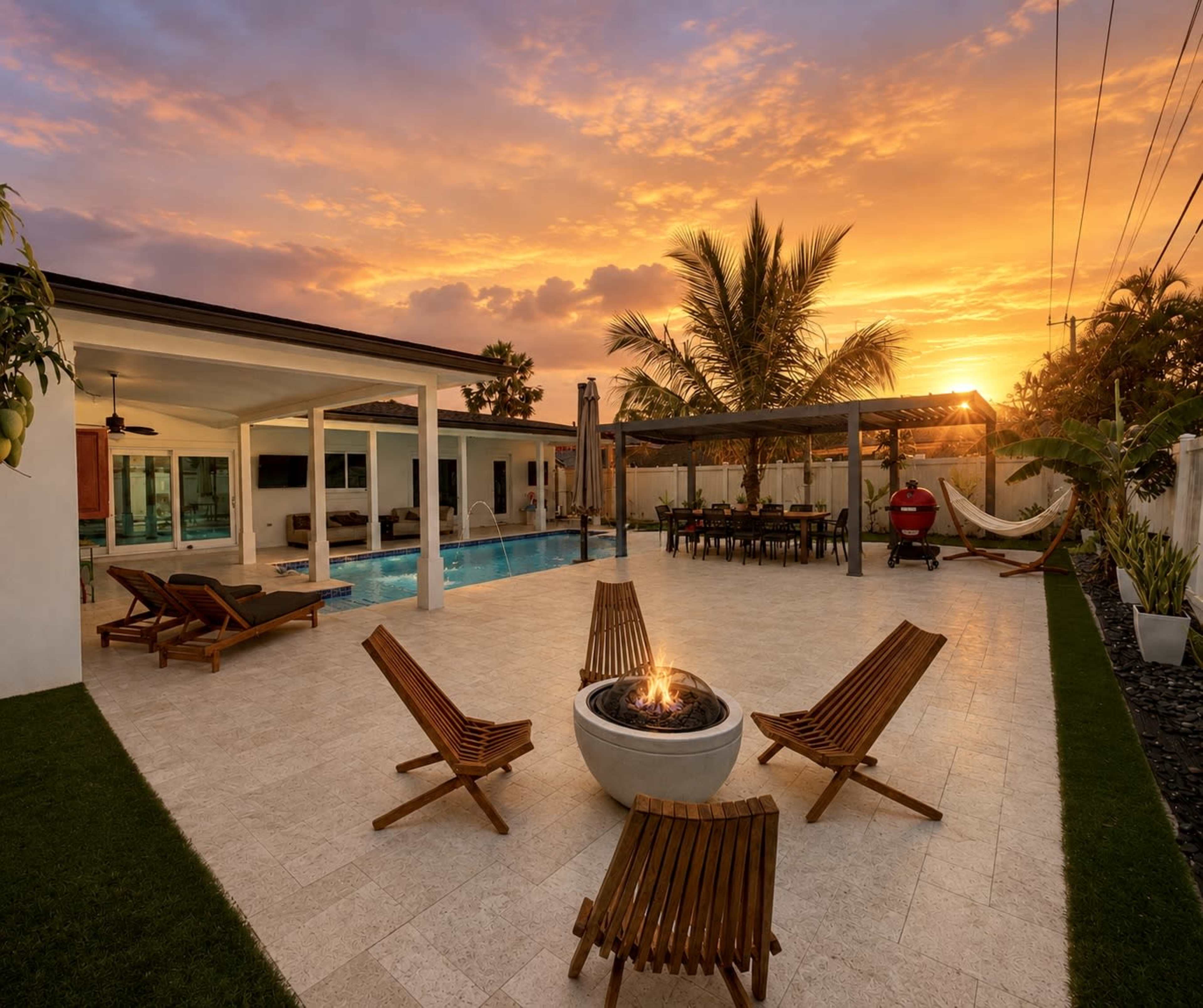 A modern outdoor area features a swimming pool, wooden lounge chairs, a firepit, and a dining space, against a backdrop of a vibrant sunset.