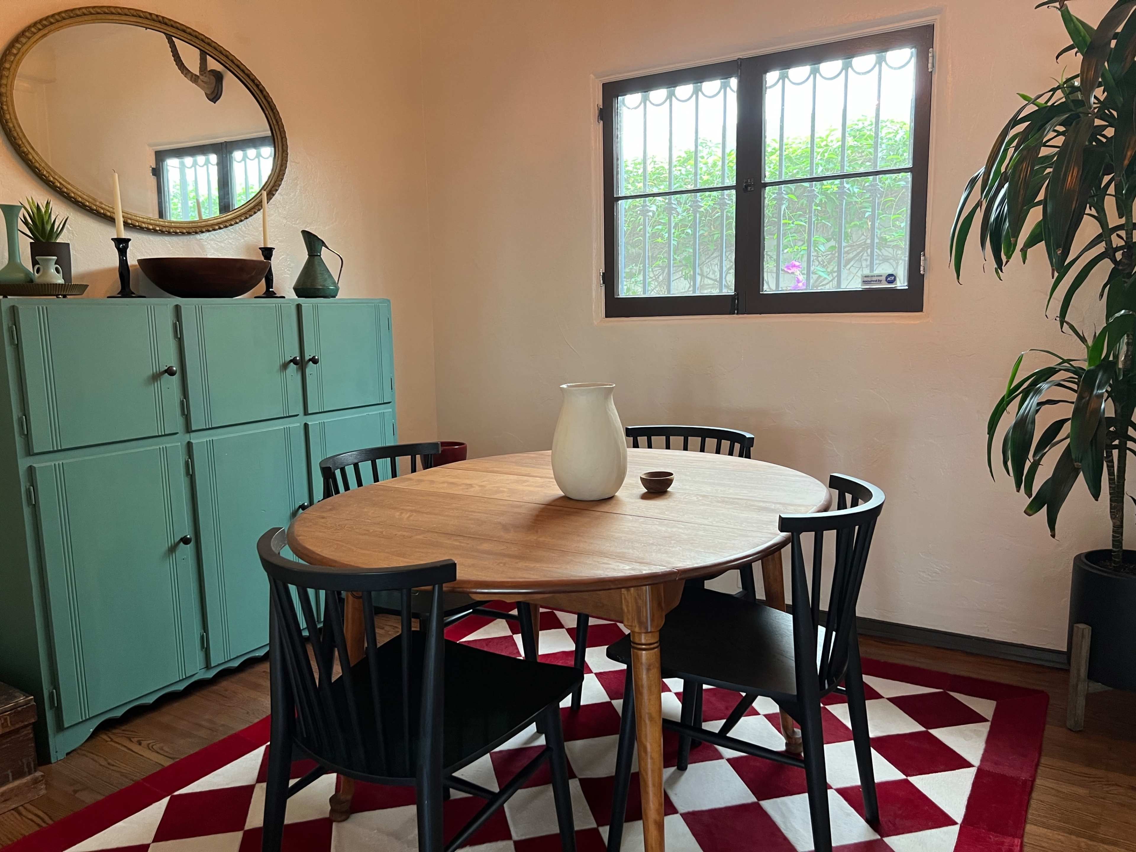 The image features a dining area with a round wooden table surrounded by black chairs, a turquoise cabinet, and a large mirror above the table, all set on a red and white patterned rug.