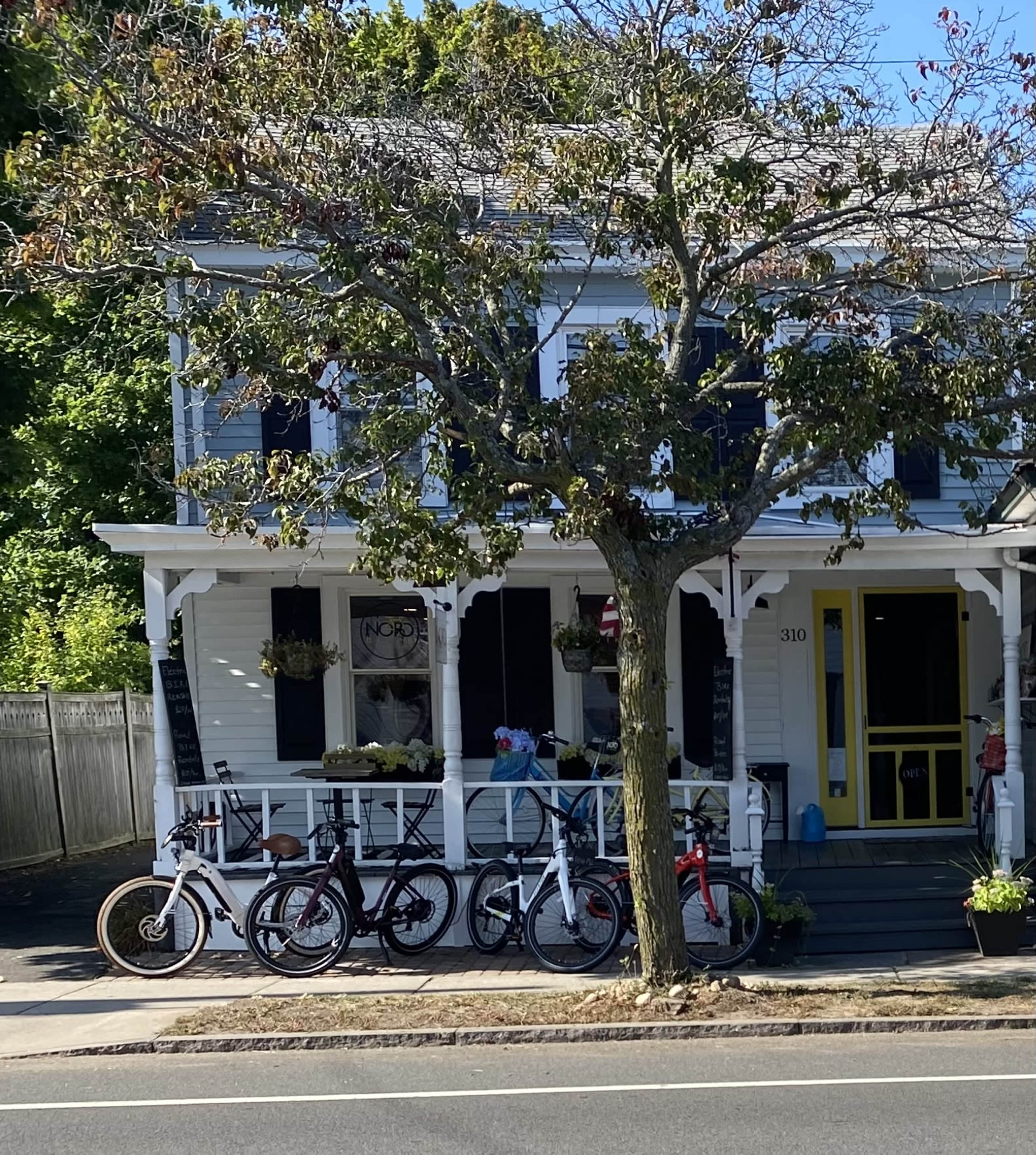 A white house with black shutters features a front porch where several bicycles are parked in front of a tree.