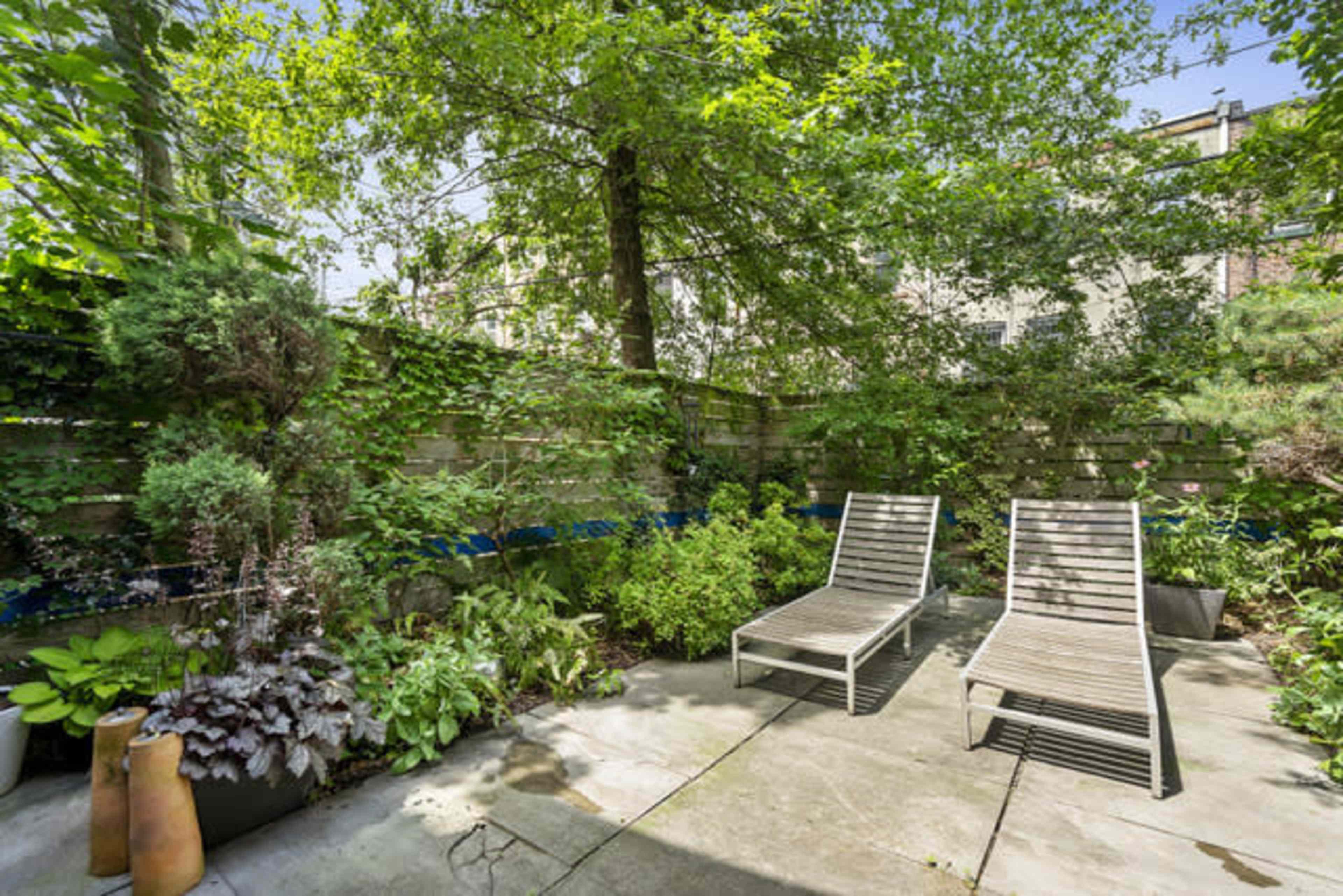 The image shows a secluded garden area with two lounge chairs surrounded by various plants and a stone wall.