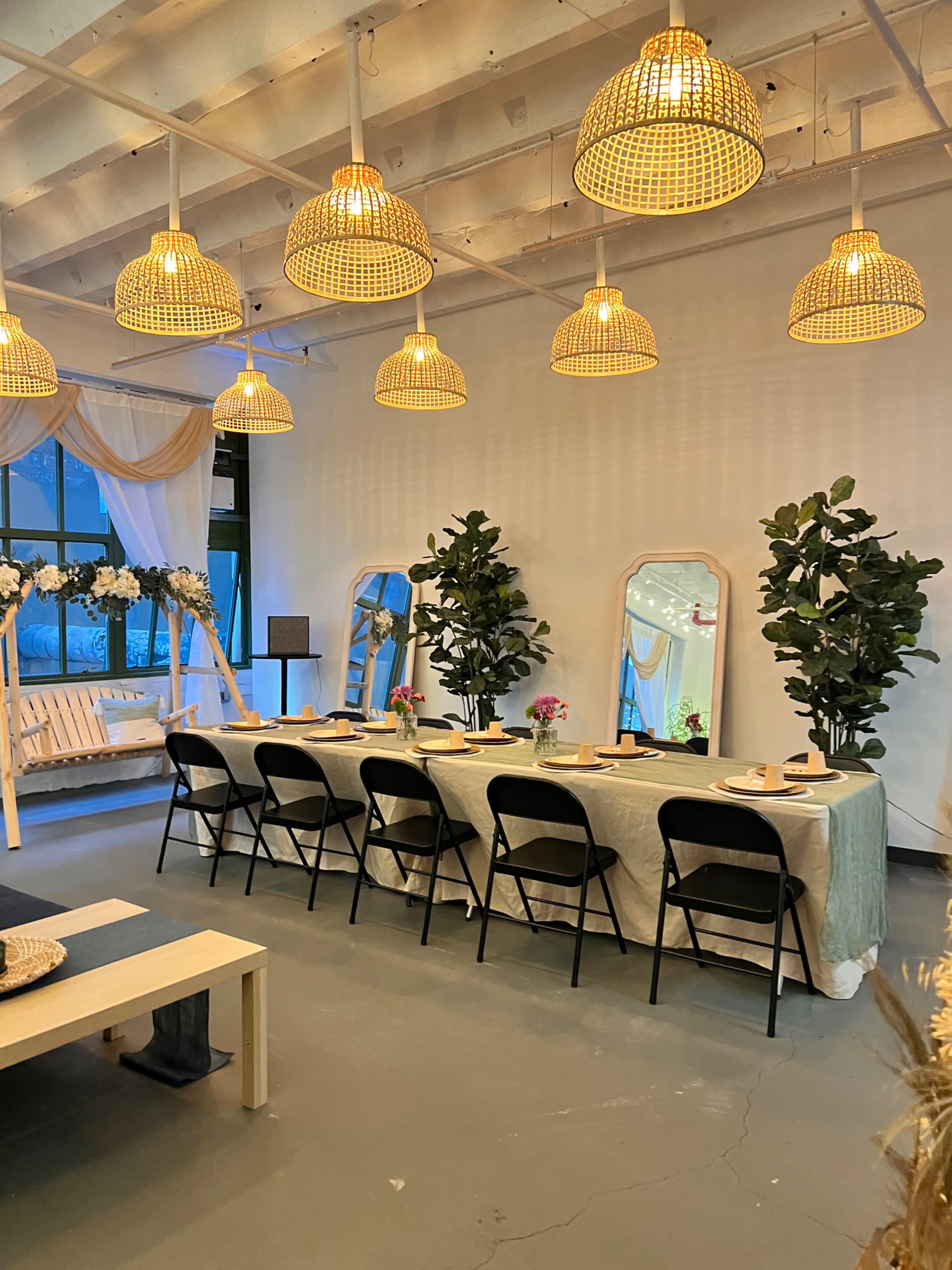 A dining area set up with a long table, black chairs, decorative mirrors, and pendant lights in a well-lit space.