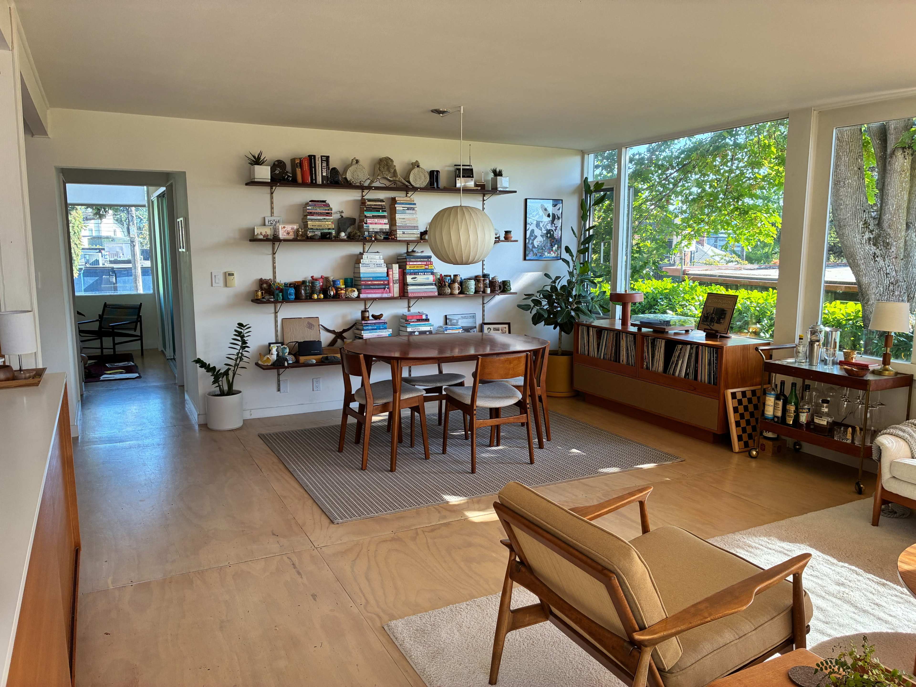 The image shows a bright, modern interior of a living space with a dining area, shelves filled with books, and large windows letting in natural light, surrounded by greenery.