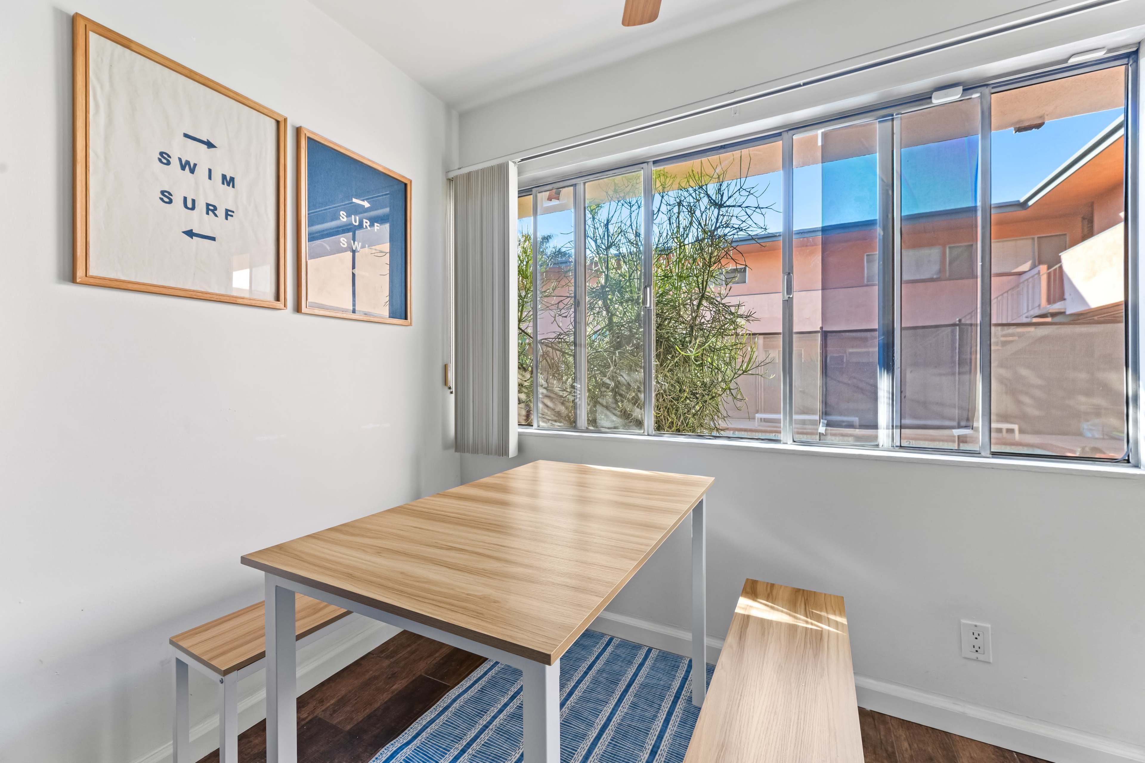 A simple dining area features a wooden table and bench, with large windows allowing natural light to enter.