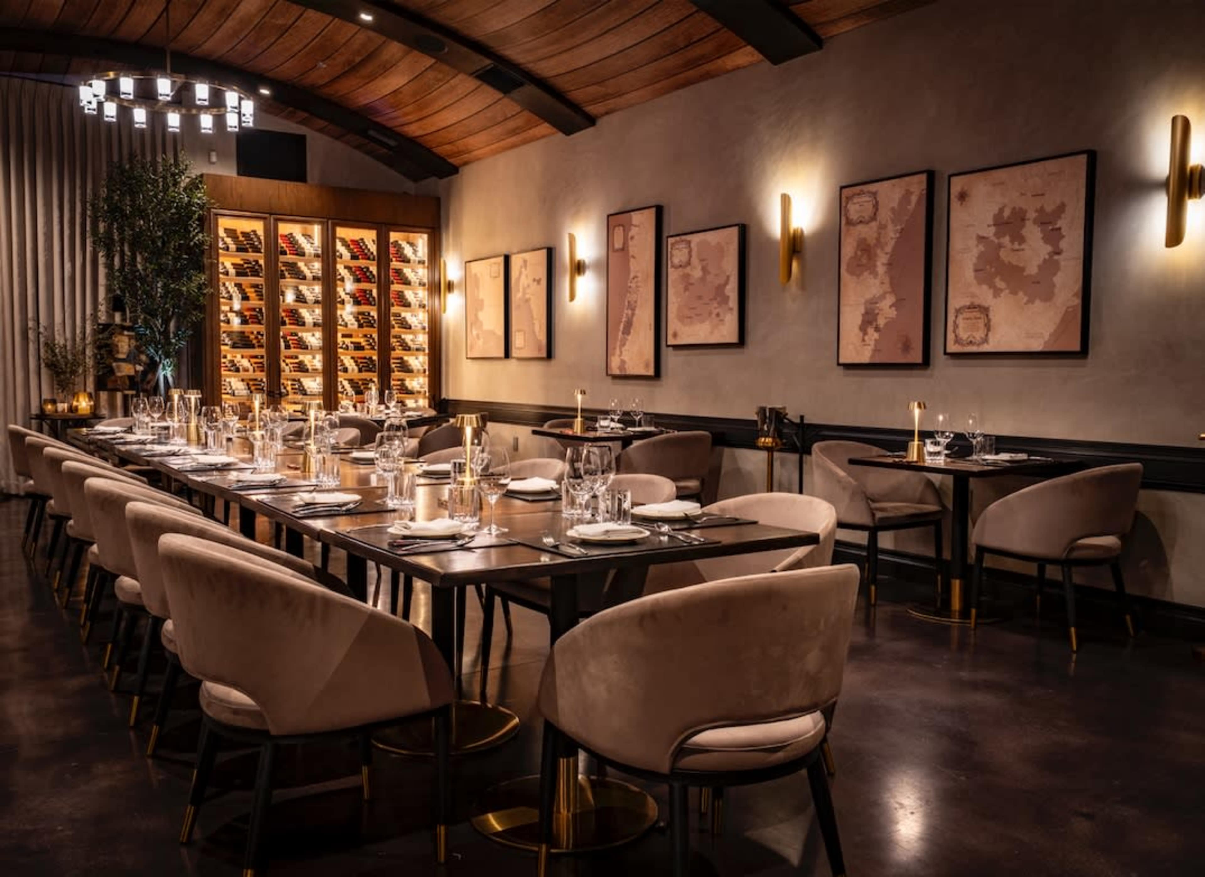 The image shows an elegantly designed restaurant featuring a long table set for dining, plush chairs, ambient lighting, and a wine display in the background.