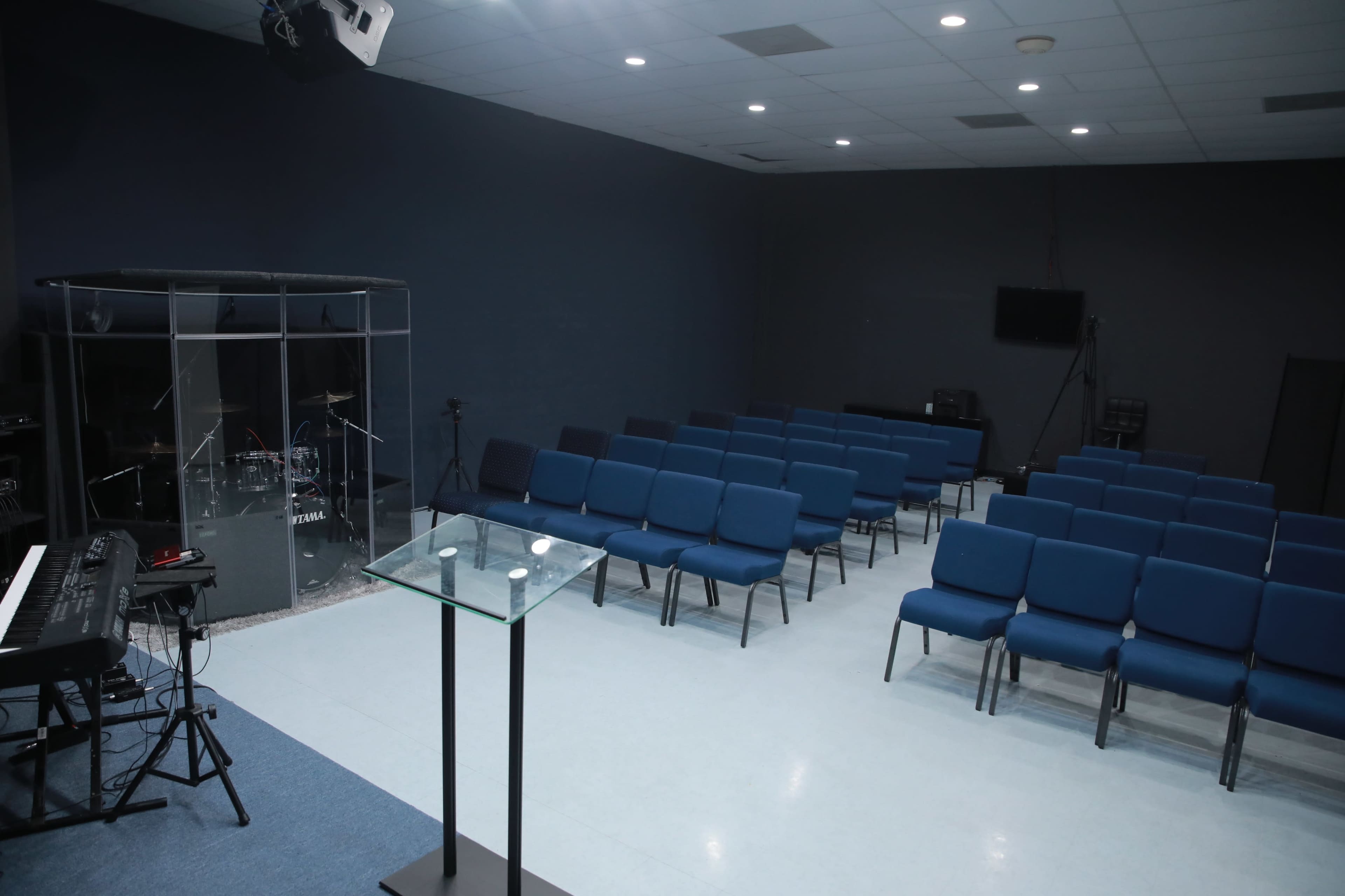 An empty room features blue chairs arranged in rows facing a glass-enclosed drum set and a transparent lectern.