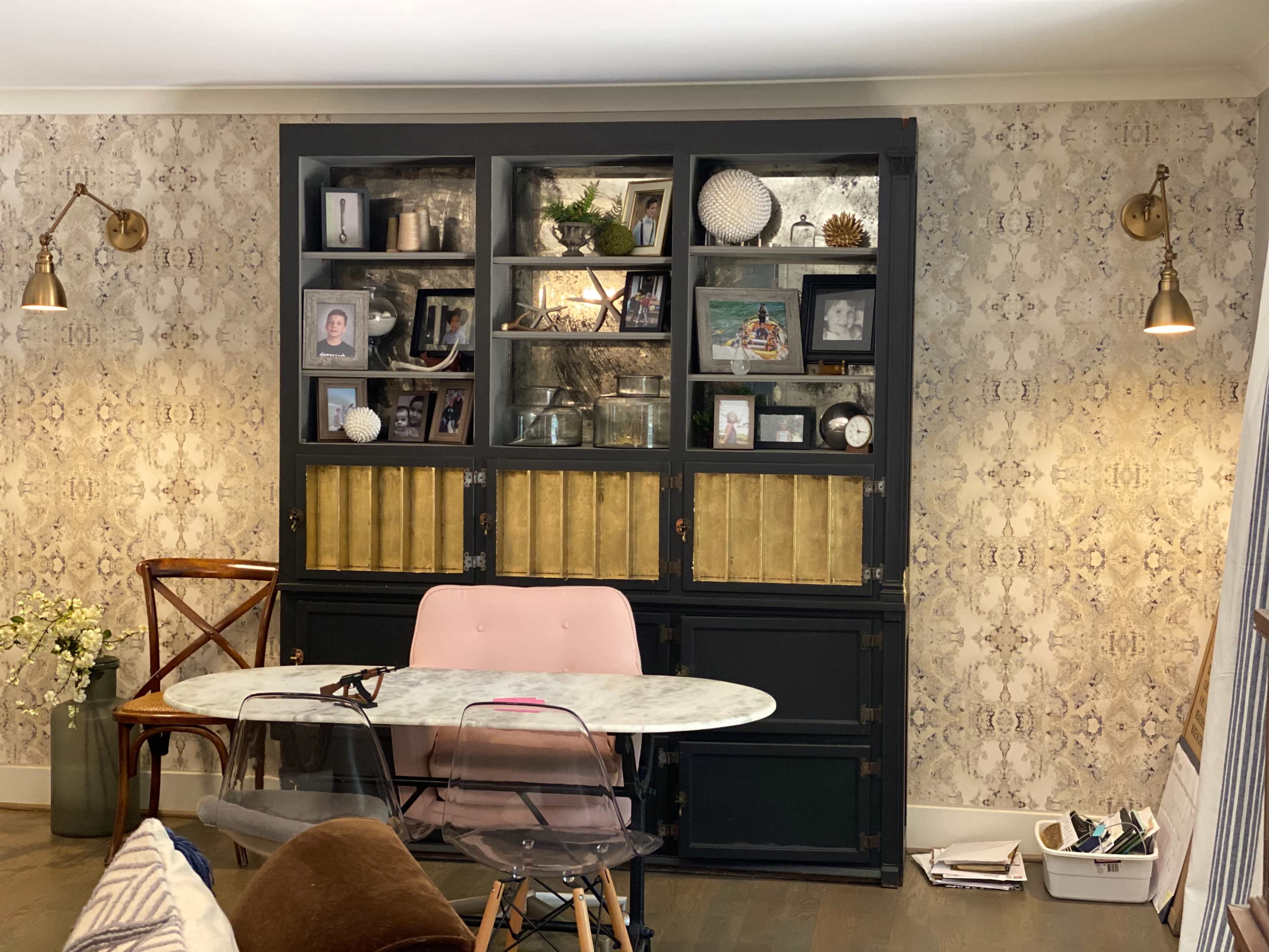 A black and gold bookshelf filled with photos and decor is placed against a patterned wall, with a round marble table and chairs in front.