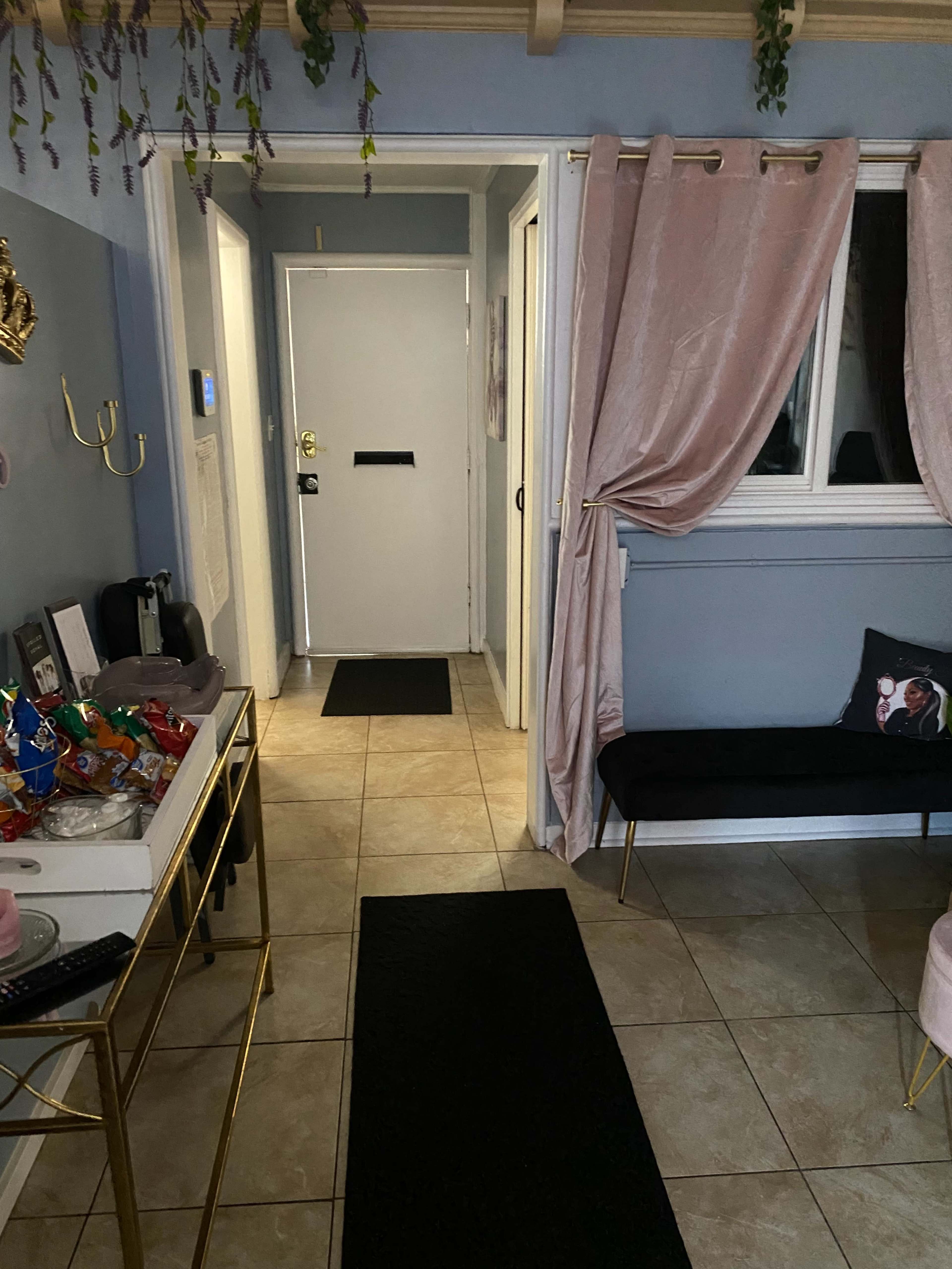 A hallway with a door at the end, flanked by a sofa, a table with snacks, and a window covered by curtains.