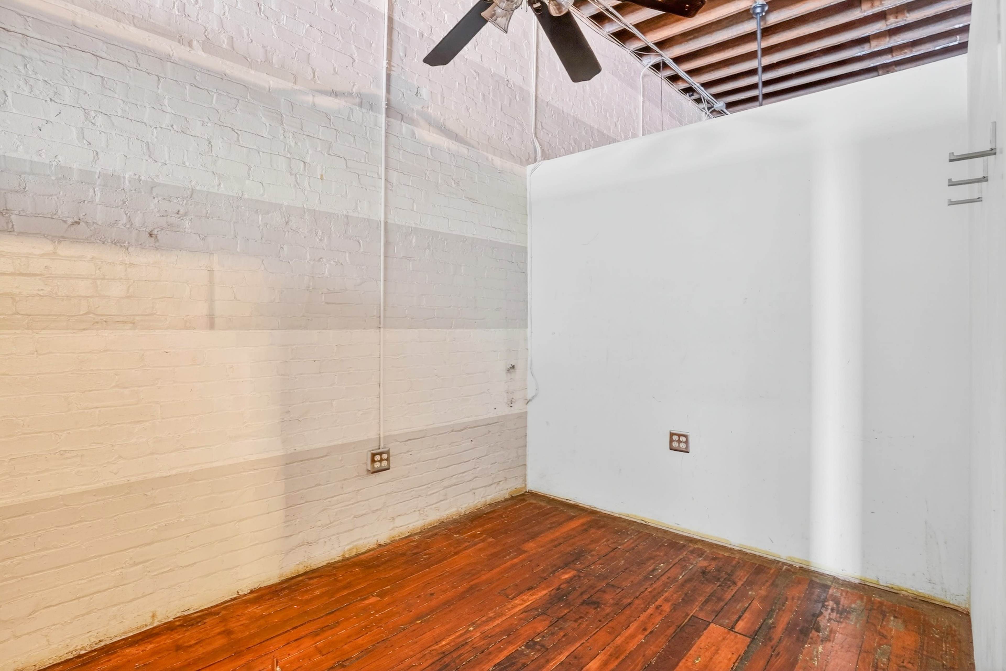 The image shows a small, empty room with exposed wooden flooring and a ceiling fan, featuring white brick walls and a sloped ceiling.