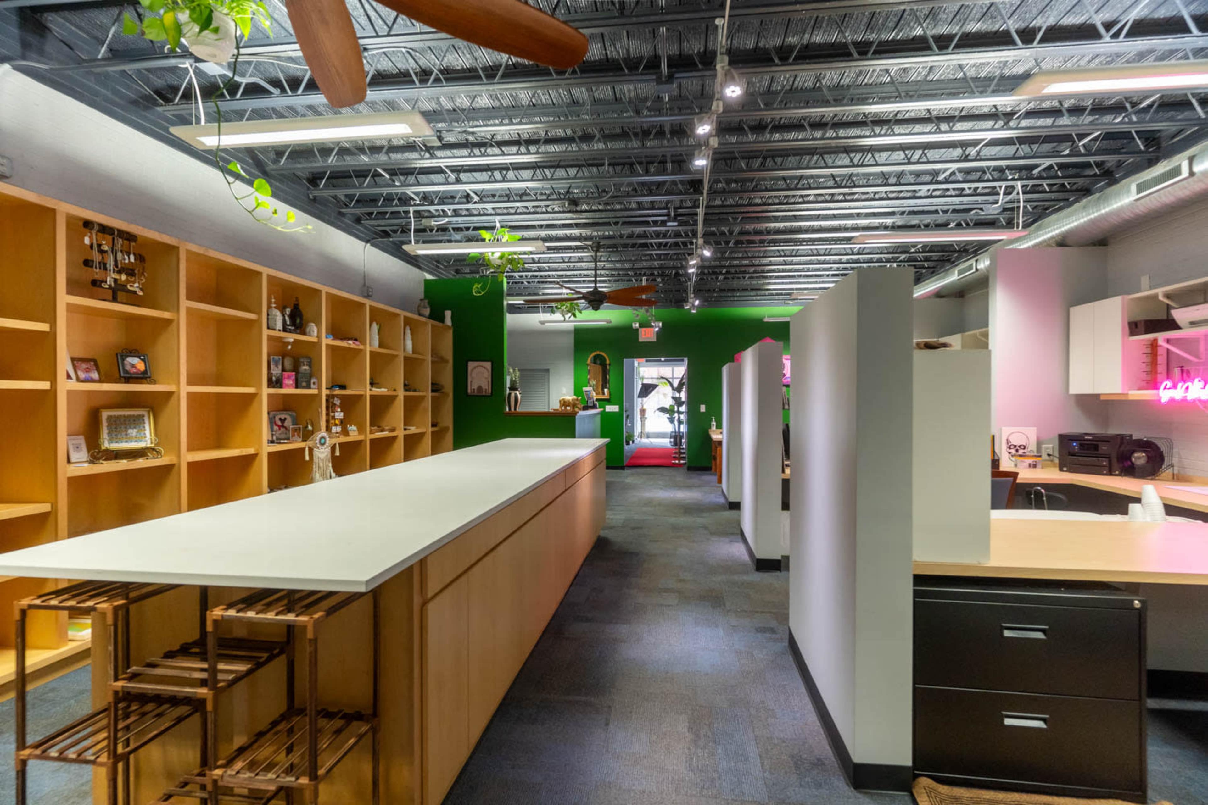A modern office space with wooden shelves on one side, a long countertop in the center, and bright green walls accented by potted plants.