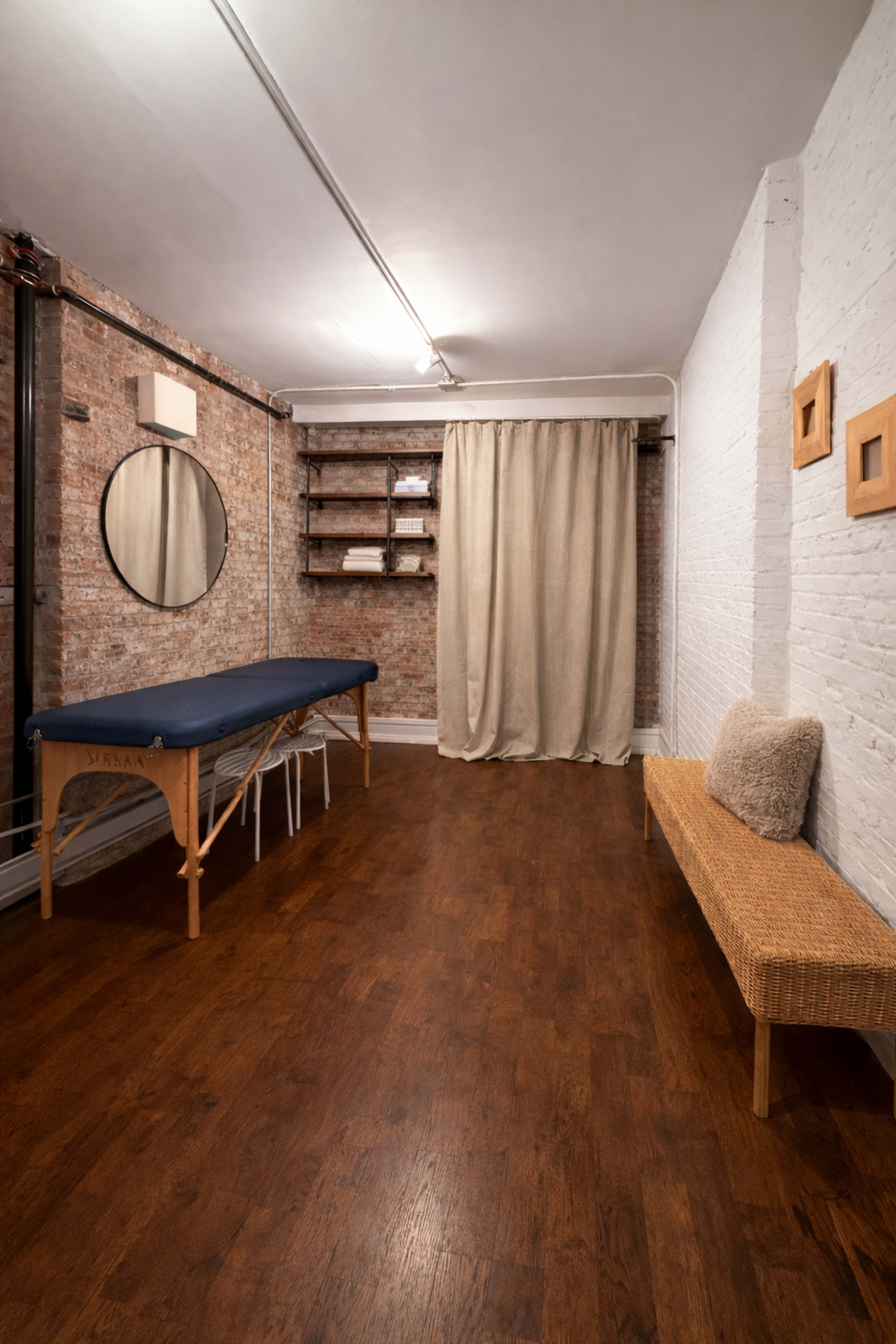 The image depicts a therapy room featuring a massage table, a bench, a round mirror on the wall, and a curtain separating the space.