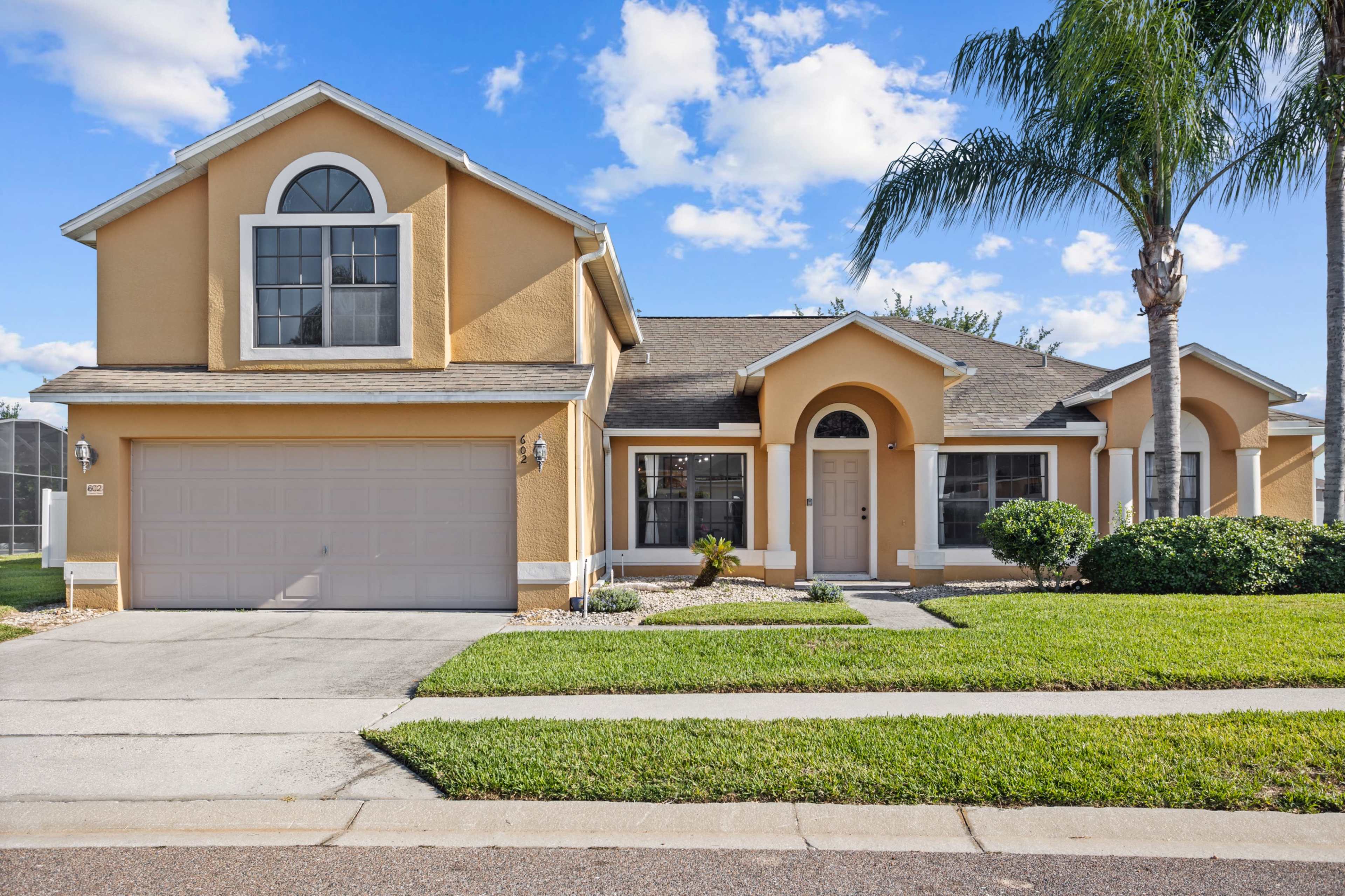 A two-story, yellow suburban house with a garage and a palm tree in the front yard.
