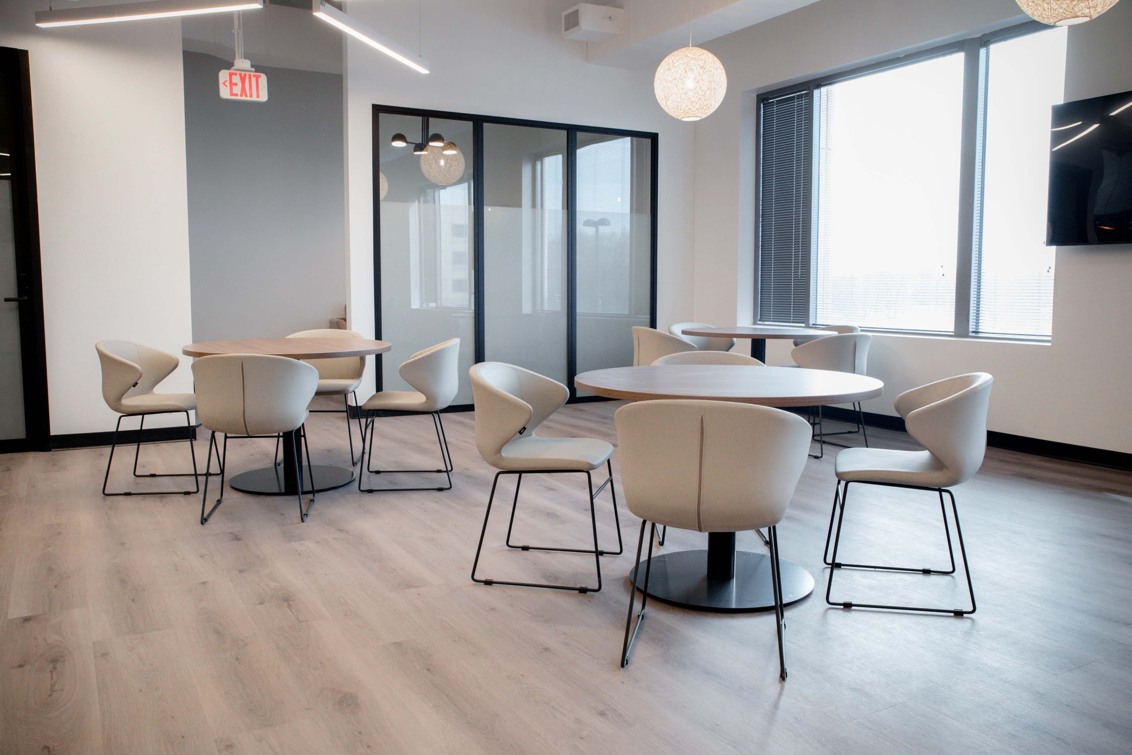 A modern office space features several round wooden tables surrounded by light-colored chairs, with large windows allowing for natural light.