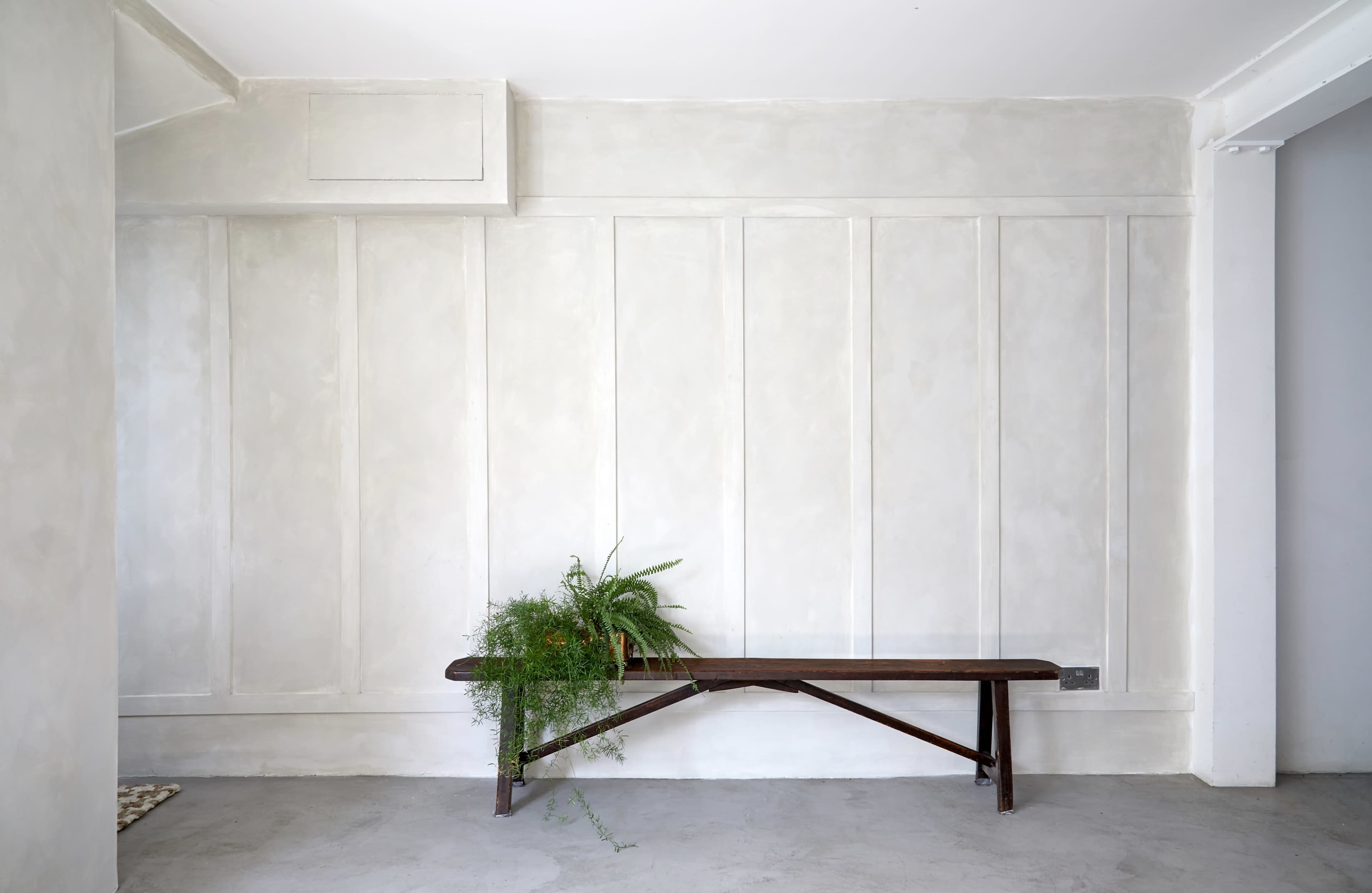 A wooden bench is placed against a textured, light-colored wall adorned with vertical paneling and a small potted plant.