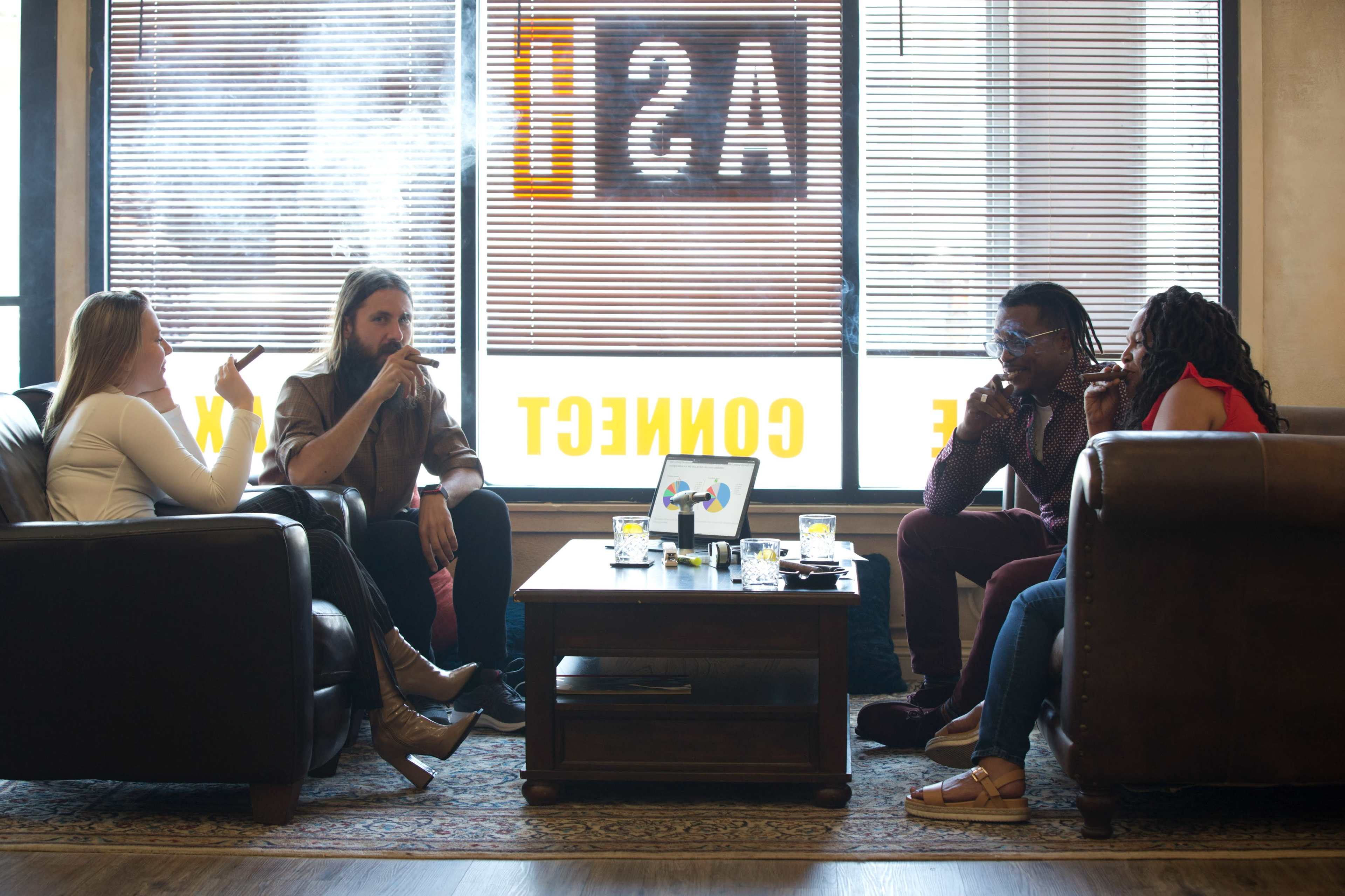 A group of four people sits on couches in a lounge, smoking and engaging in conversation around a coffee table.