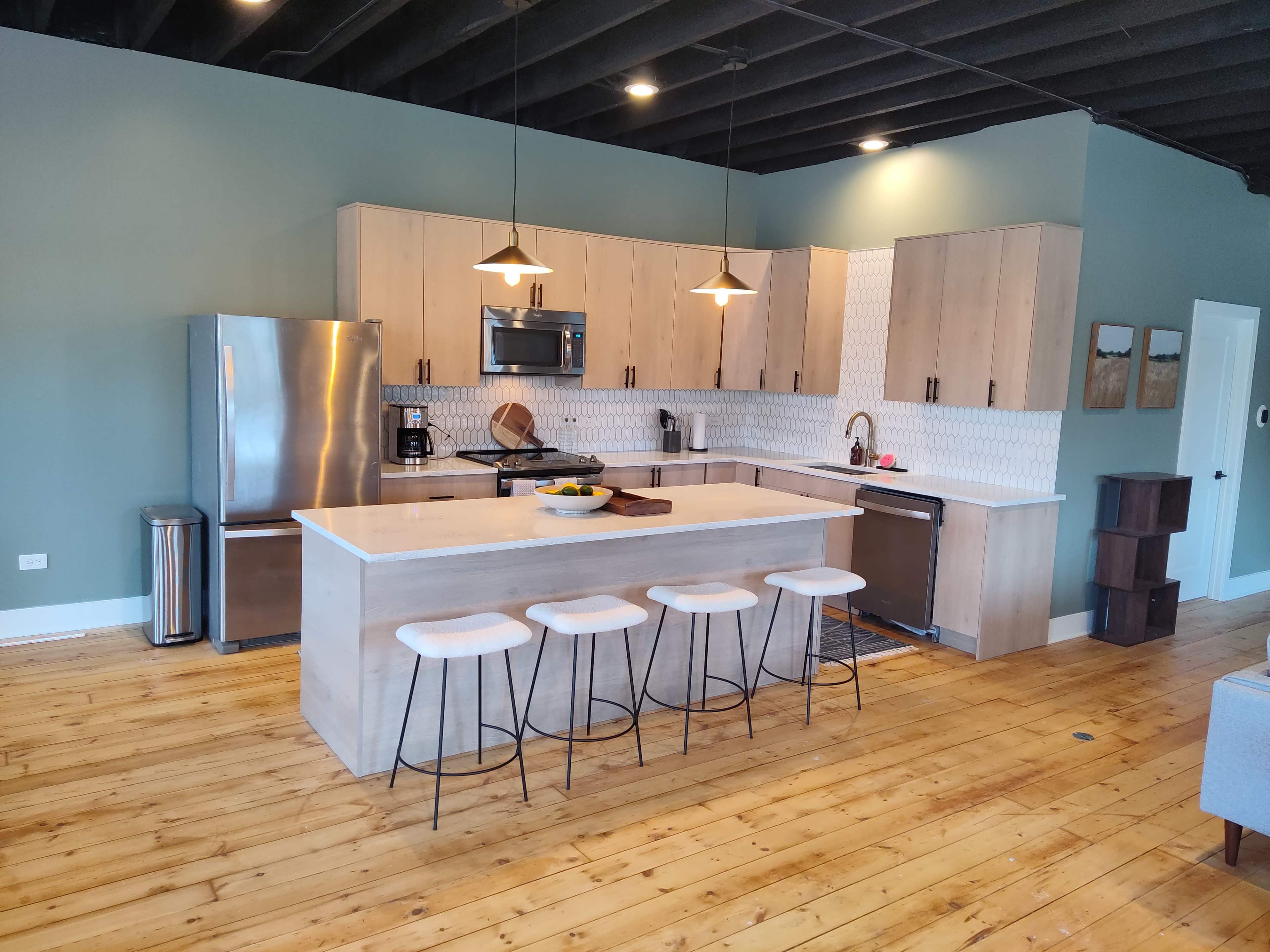 A modern kitchen featuring light wood cabinets, a large island with seating, stainless steel appliances, and a green accent wall.