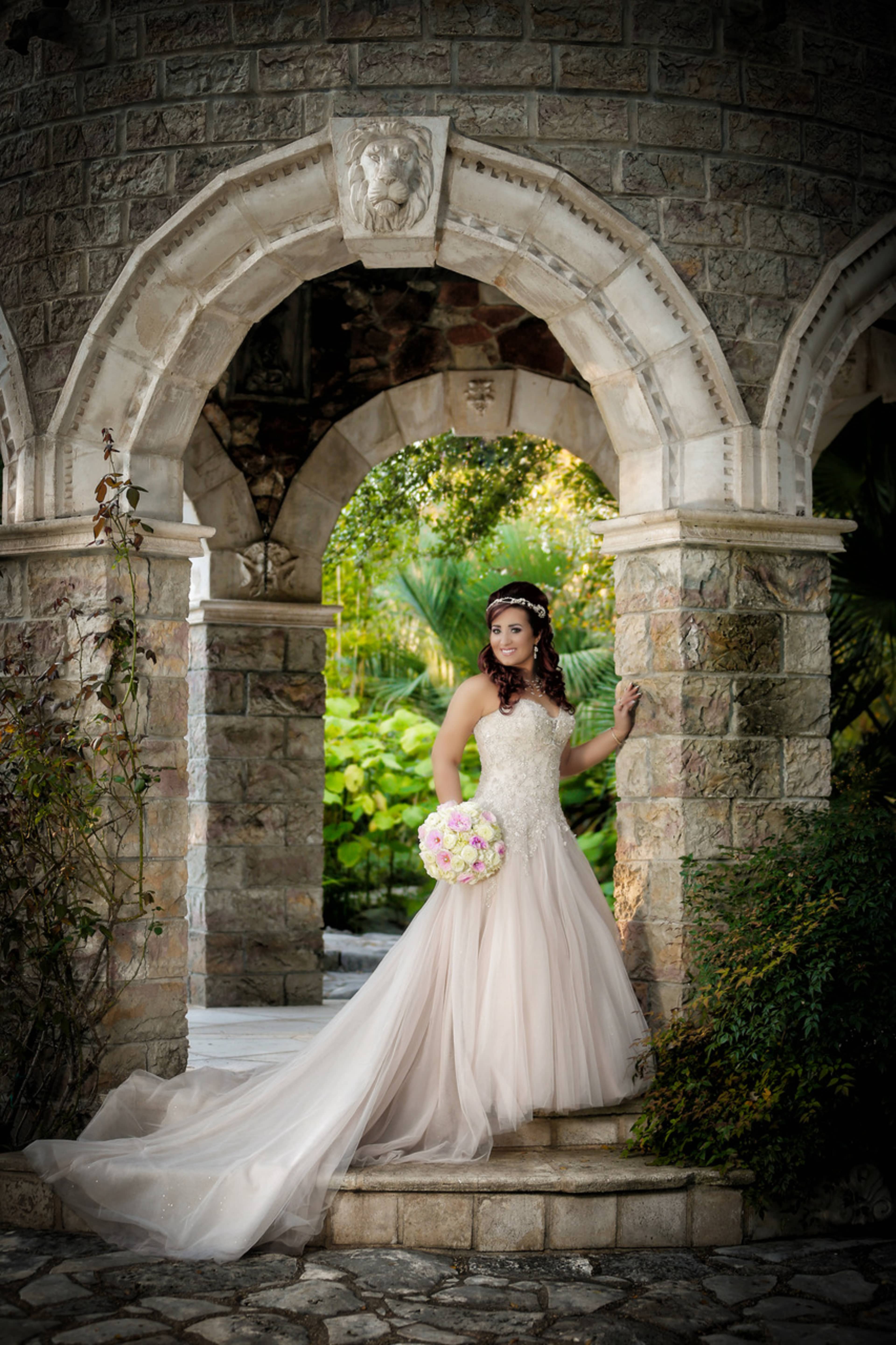 A bride in a flowing gown poses under an ornate stone archway while holding a bouquet of flowers.
