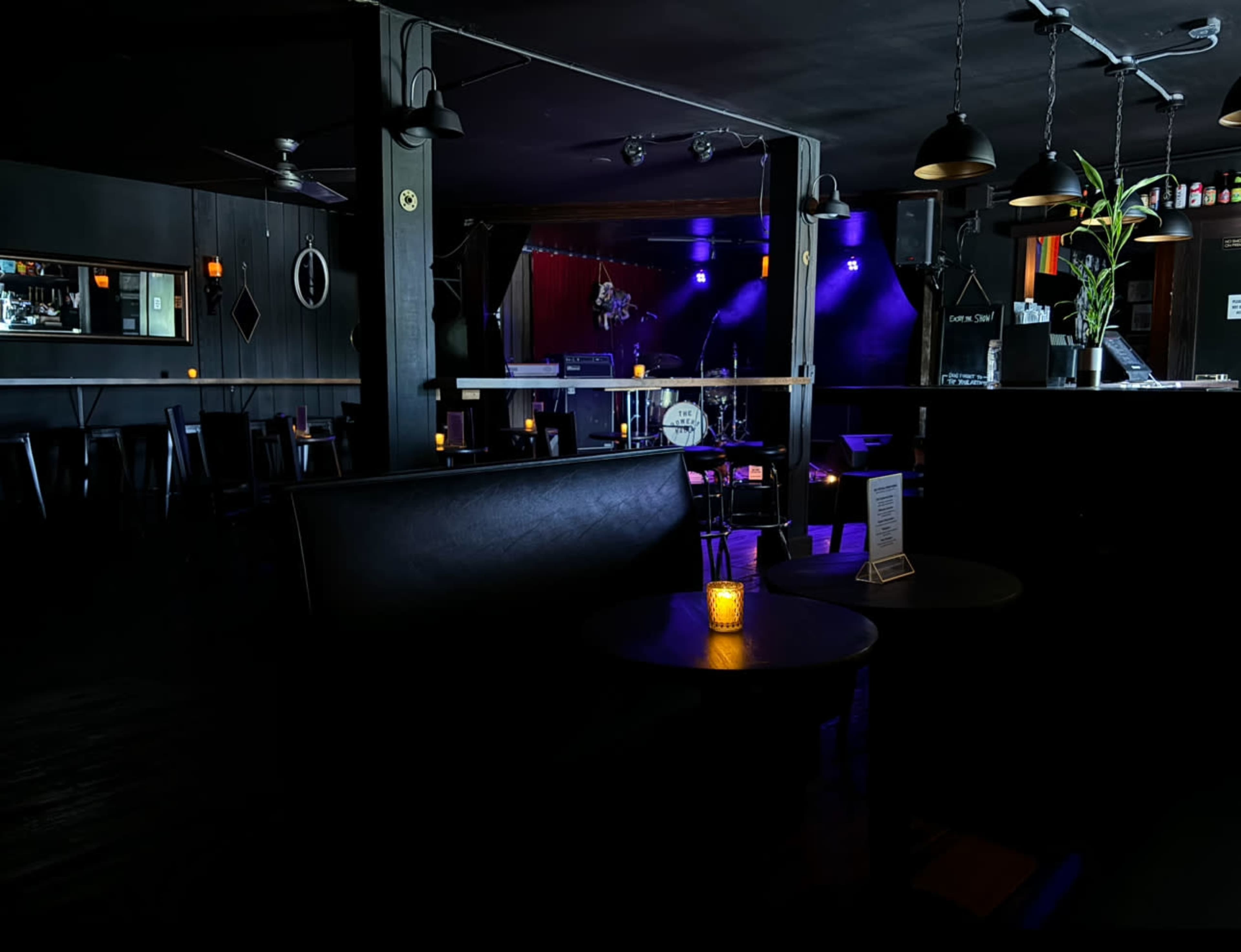 The image shows a dimly lit bar with empty tables, a stage in the background, and colorful lights.