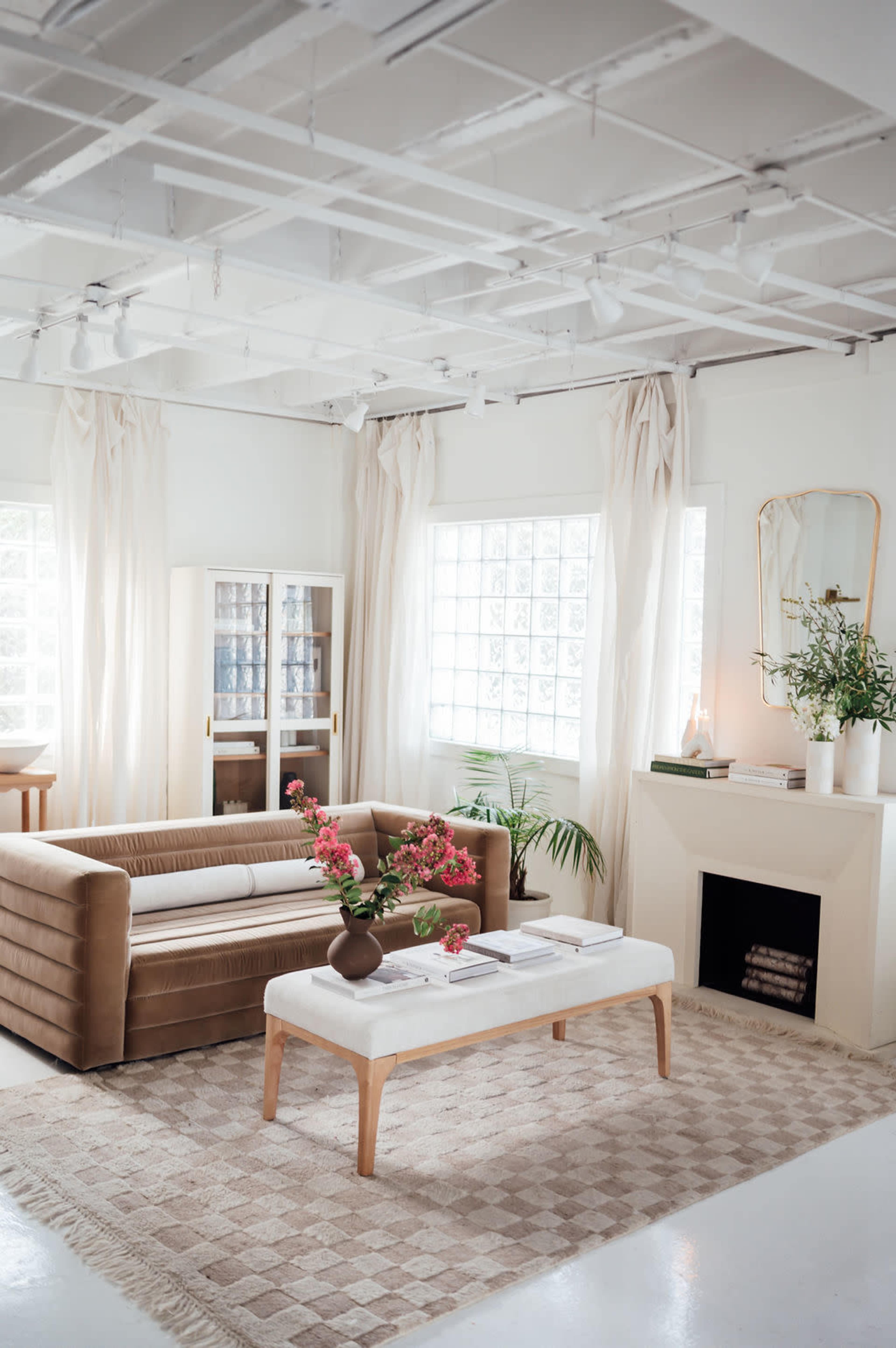 A modern living room features a brown sofa, a light-colored coffee table, and a decorative plant beside a window with sheer curtains.