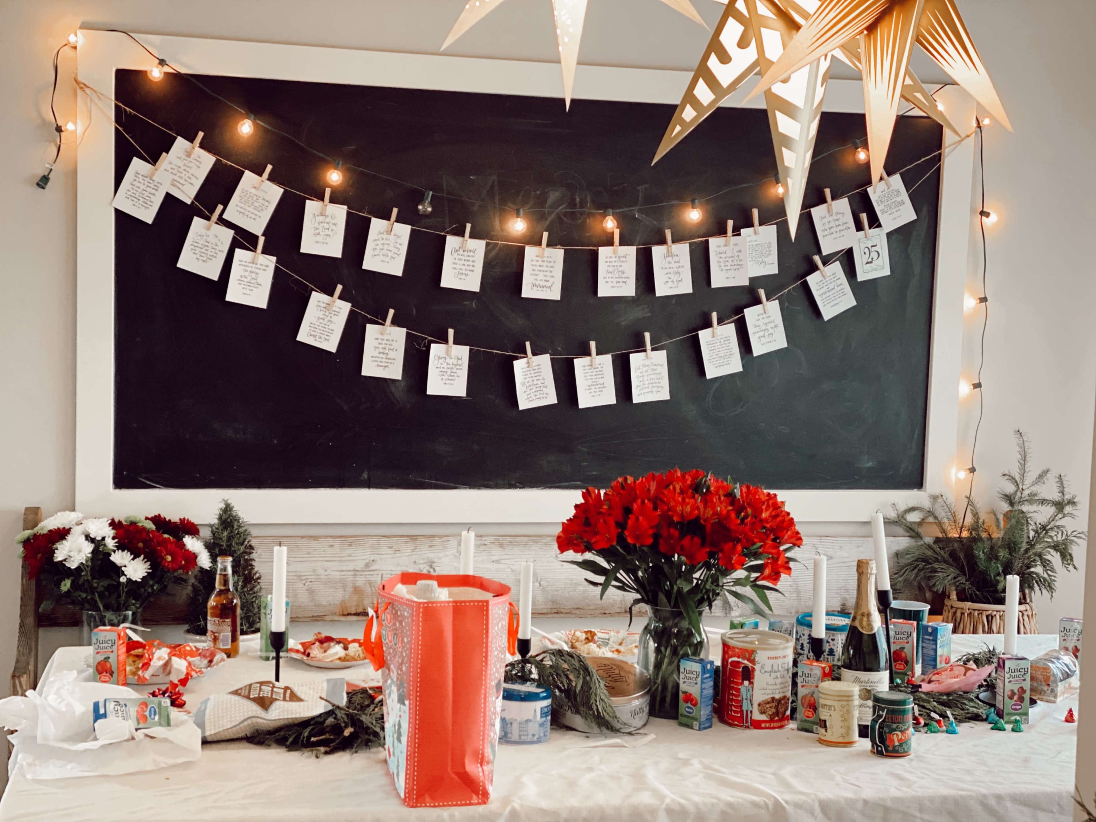 A table is decorated with red flowers, assorted food and drinks, and a string of notes hung on a chalkboard wall with fairy lights above.