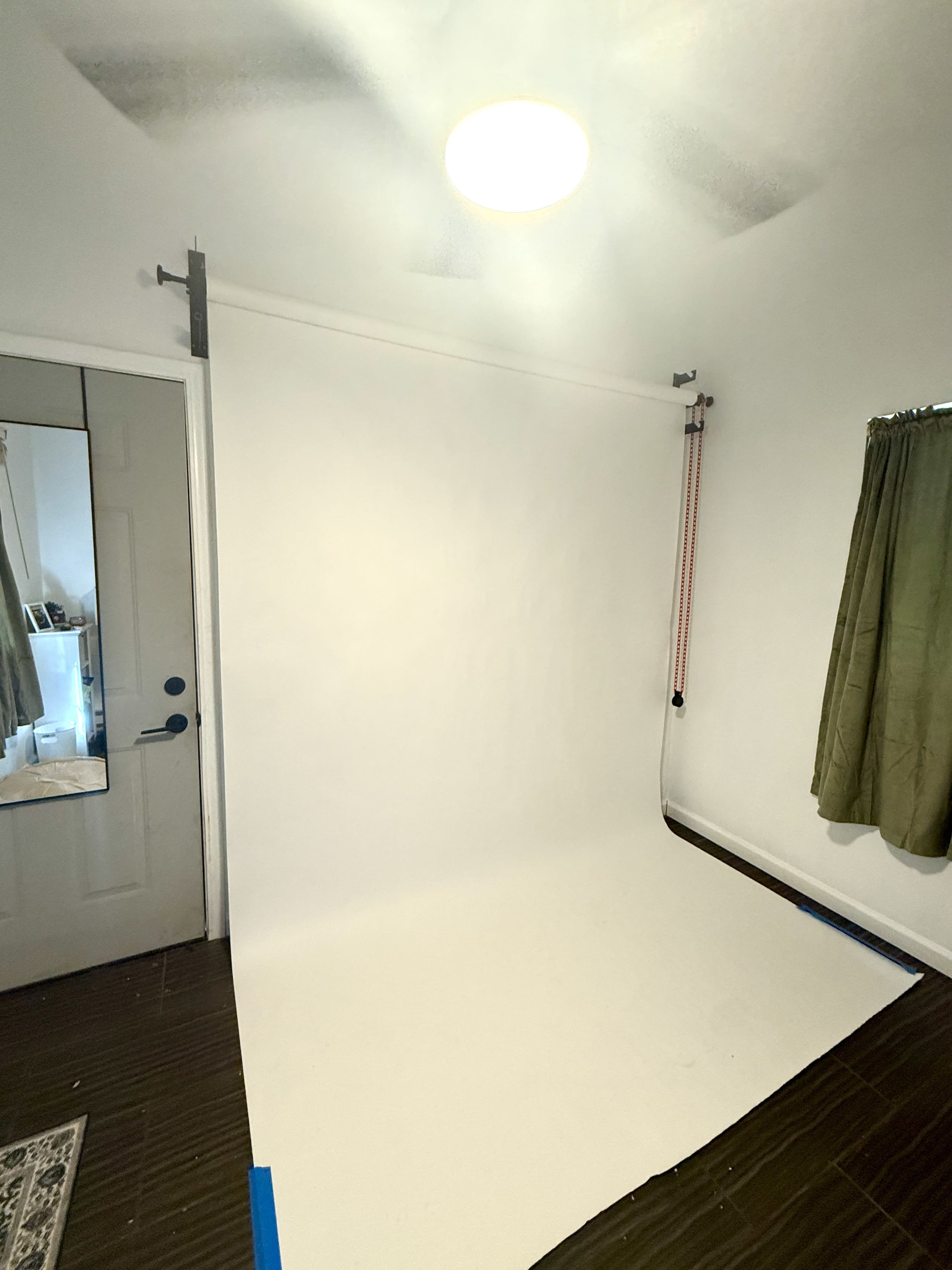 The image shows a minimalist studio setup with a white backdrop, a door, and a window covered with a green curtain.