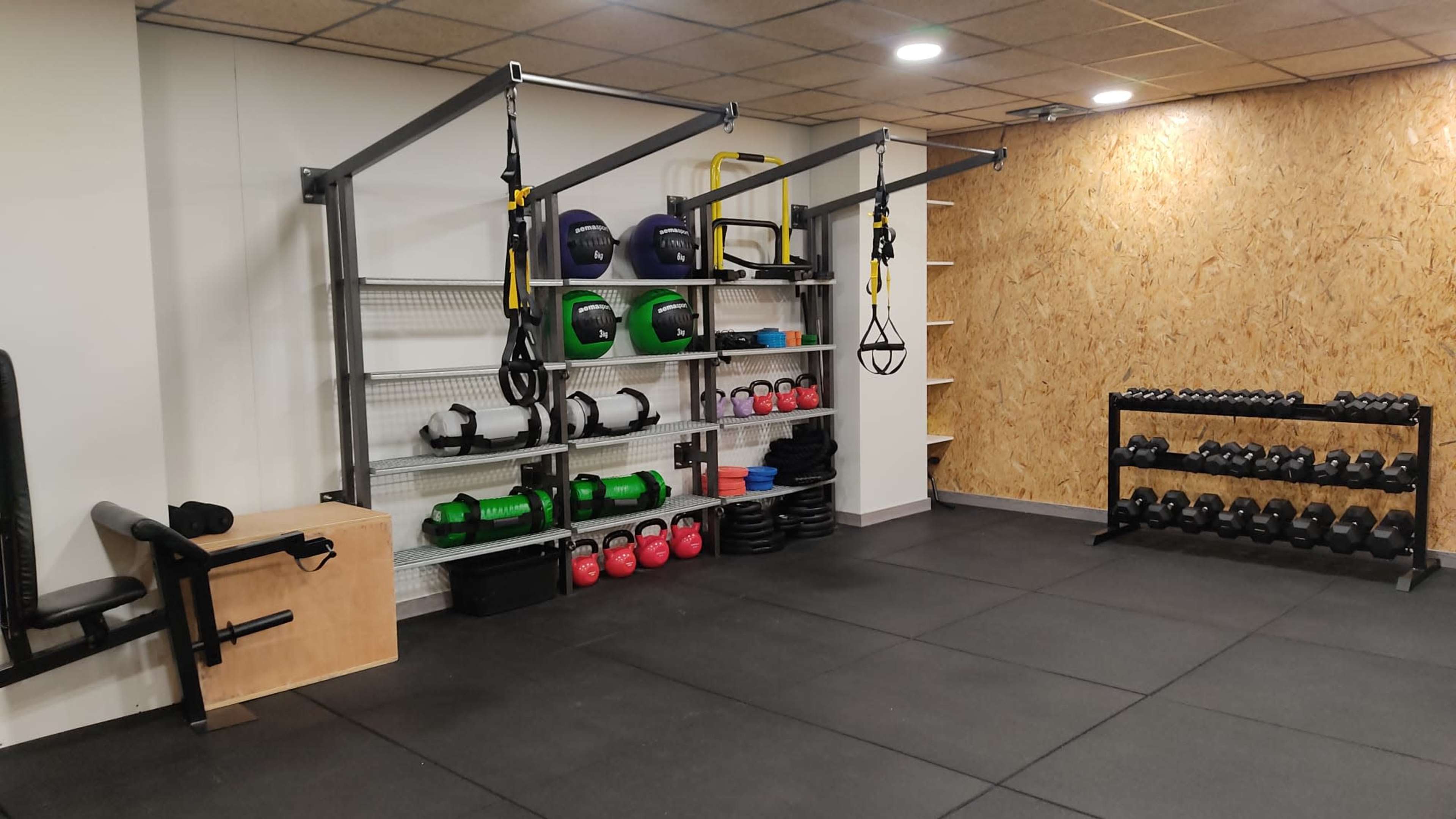 The image shows a gym interior with workout equipment, including kettlebells, dumbbells, and TRX straps organized on shelves and a rubber flooring surface.