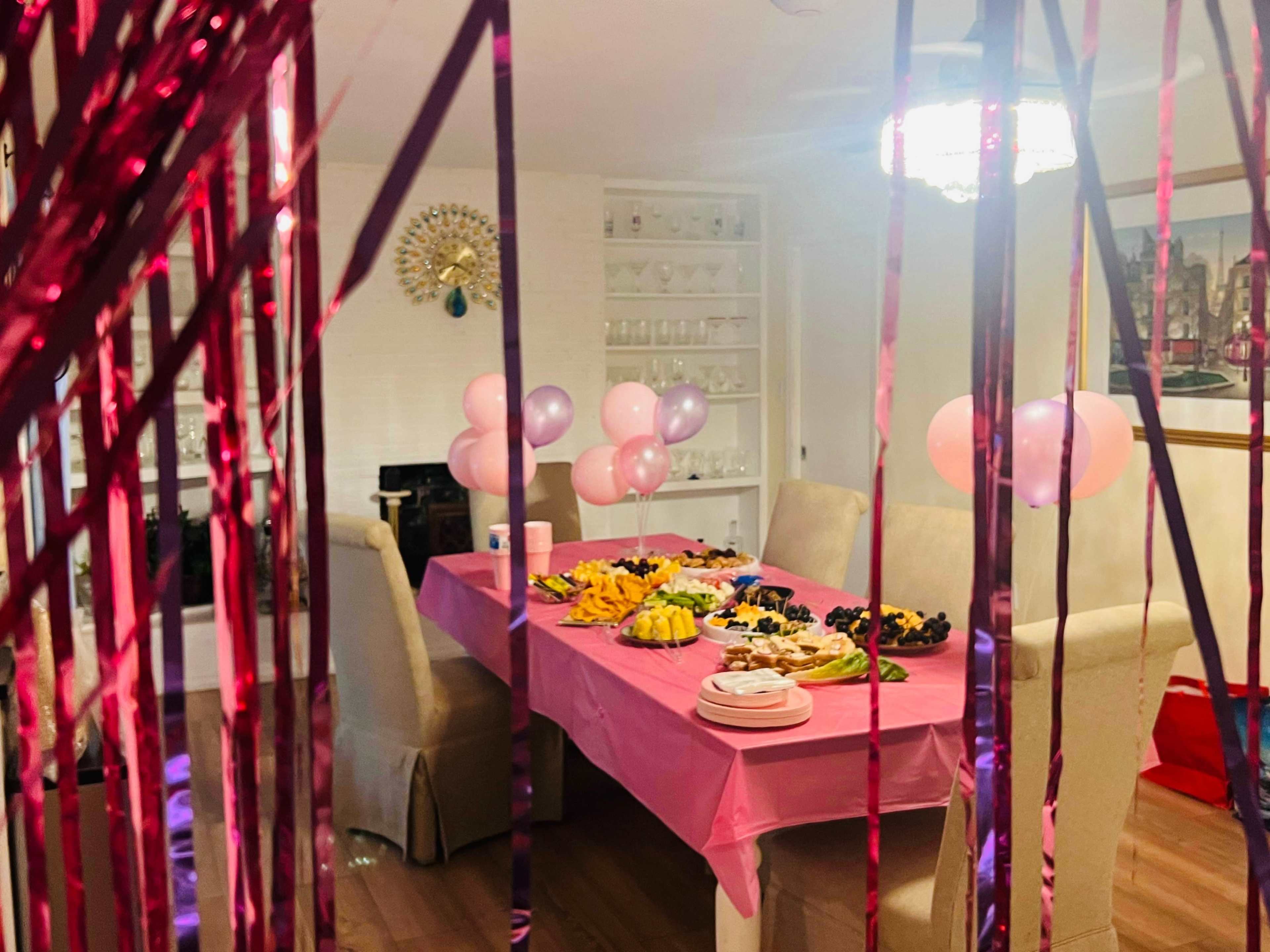A dining table covered with a pink tablecloth is filled with various fruits and pastries, surrounded by balloons and decorative streamers.