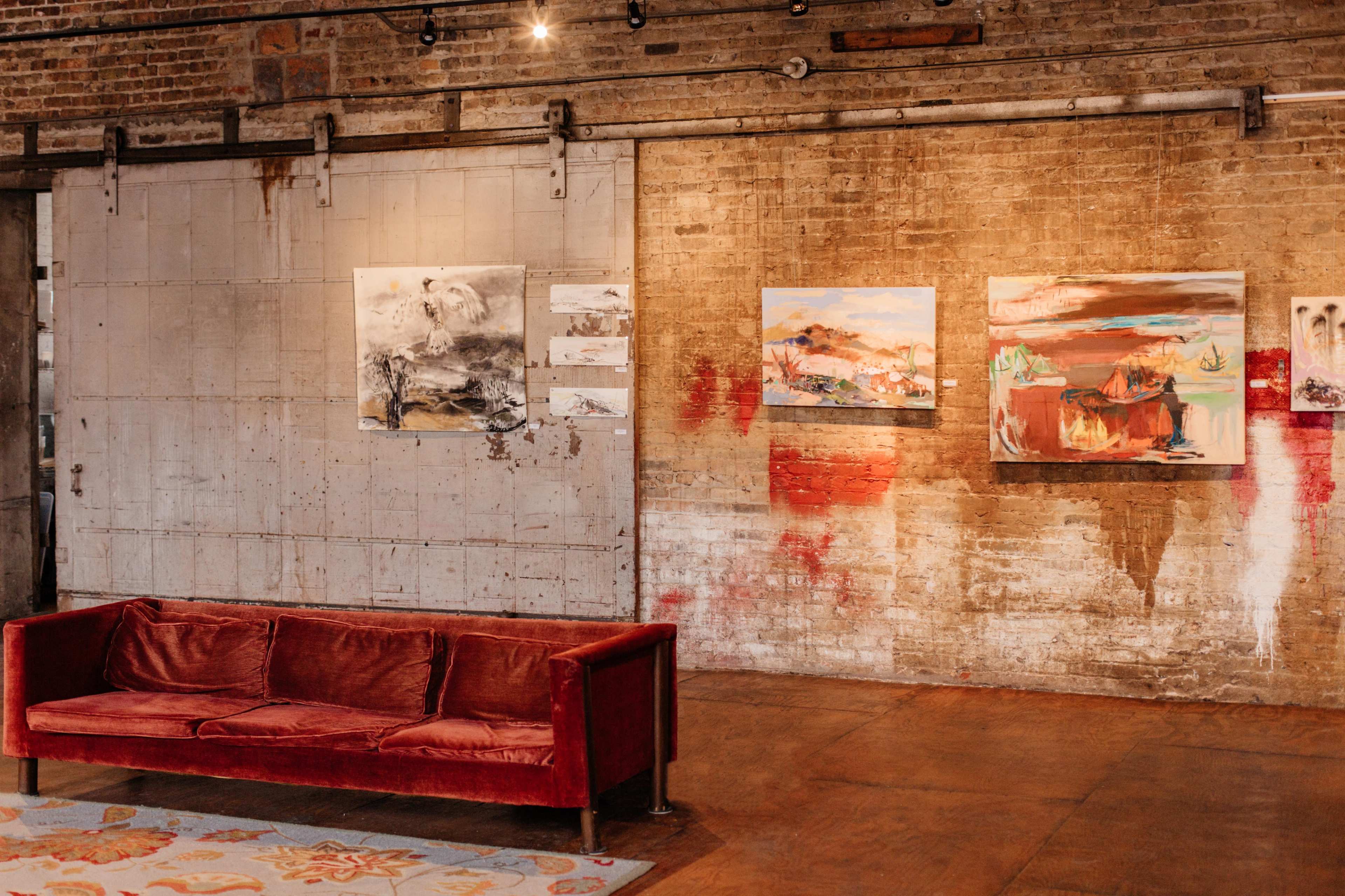 A modern art gallery featuring a red velvet sofa against a brick wall adorned with abstract paintings.