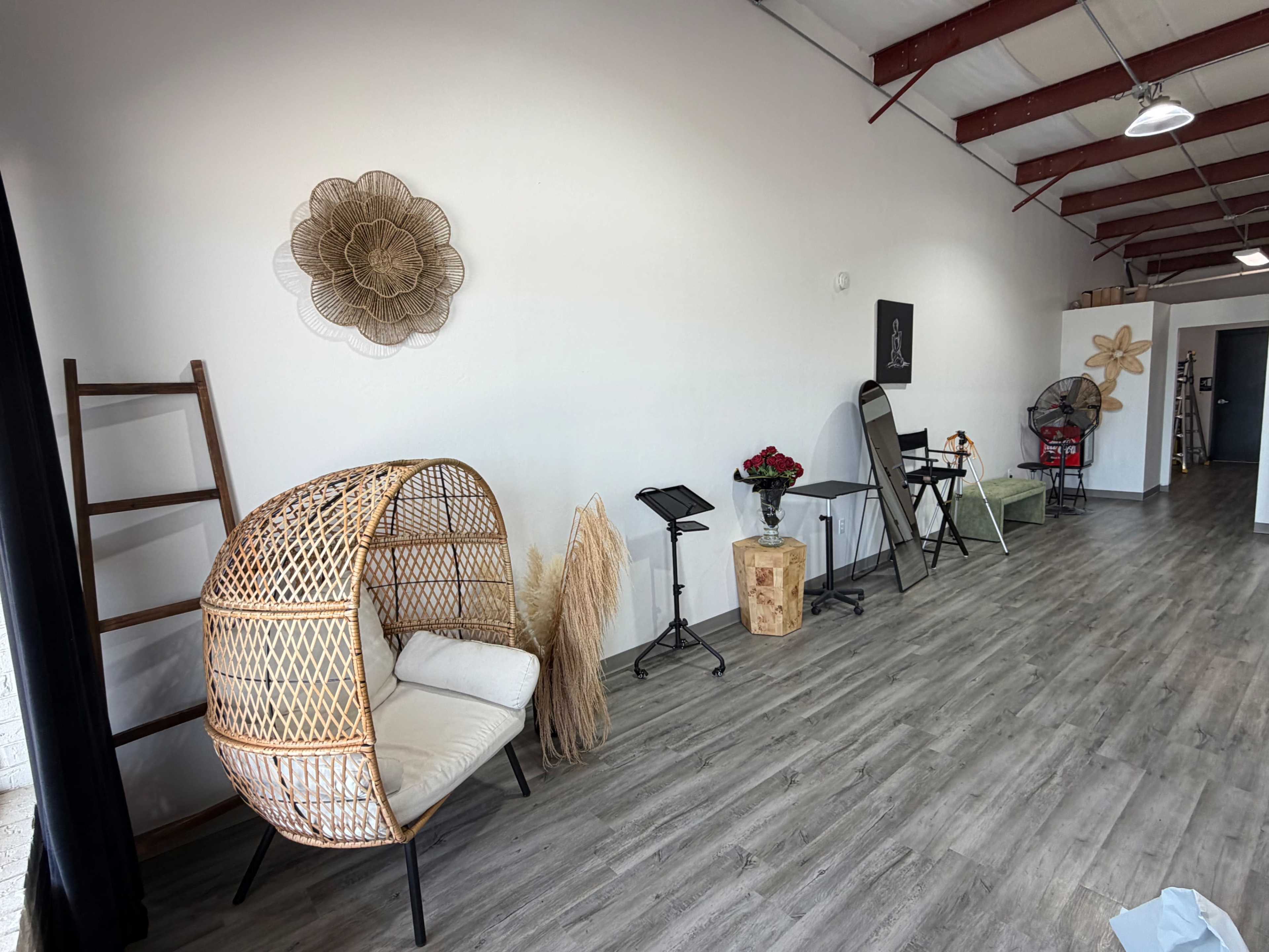 The image shows a spacious studio with wooden flooring, featuring a decorative rattan chair, a ladder, a plant stand, and various furniture arrangements along the walls.