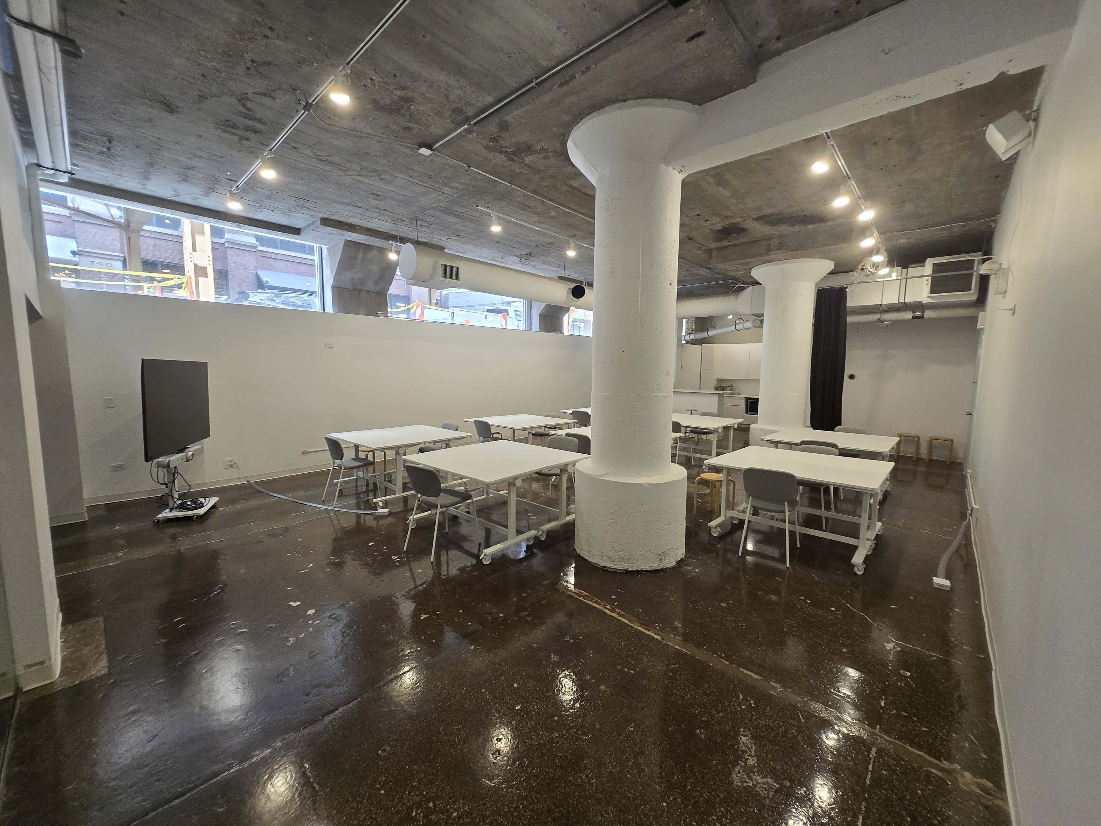 The image shows a modern, empty classroom or meeting space with several tables and chairs arranged, white walls, and exposed concrete ceilings and pillars.