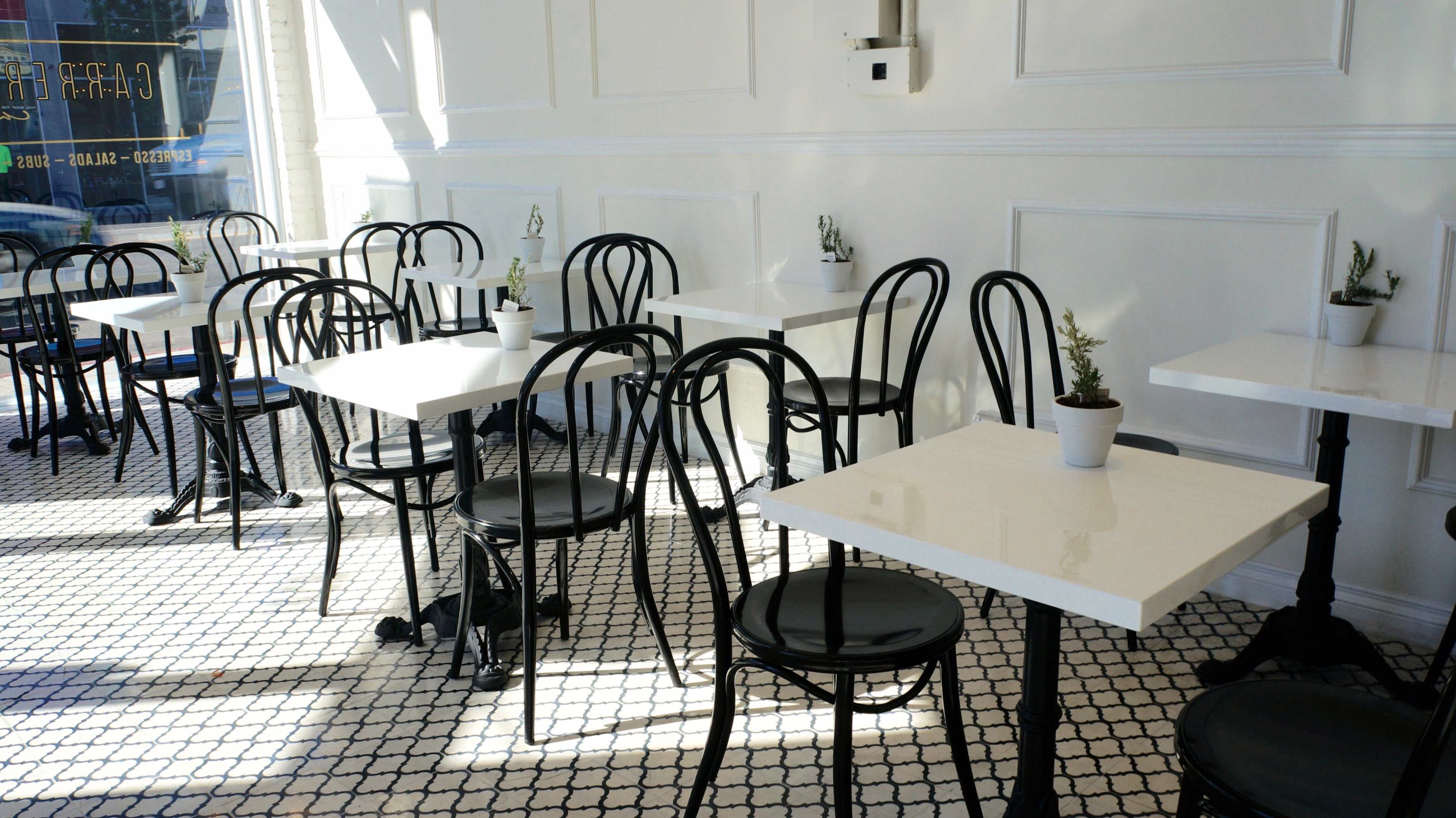 An empty café with black chairs and white tables arranged on a tiled floor, illuminated by natural sunlight.