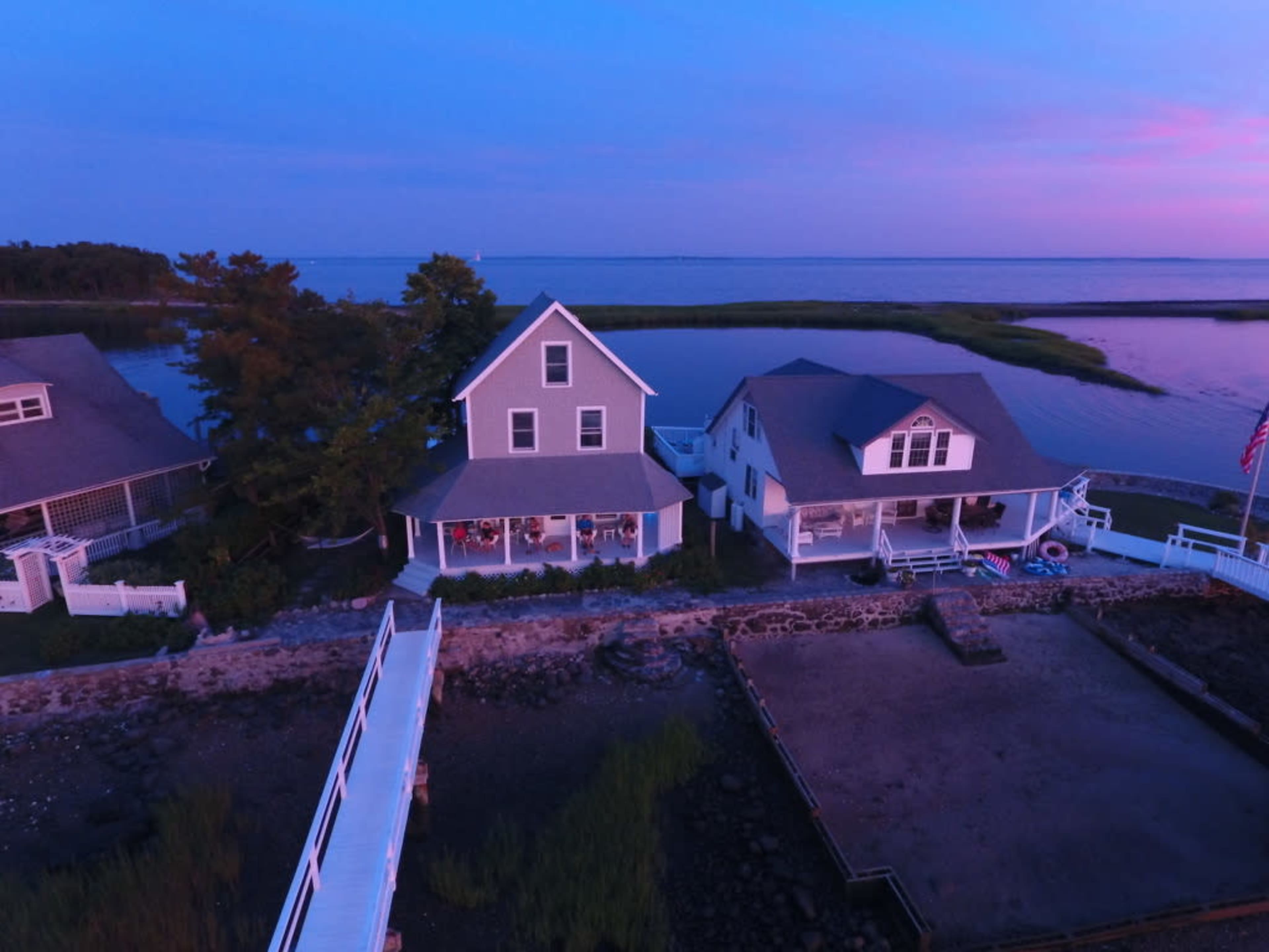The image shows a waterfront scene featuring two houses near a body of water at twilight, with a wooden dock extending towards the shore.