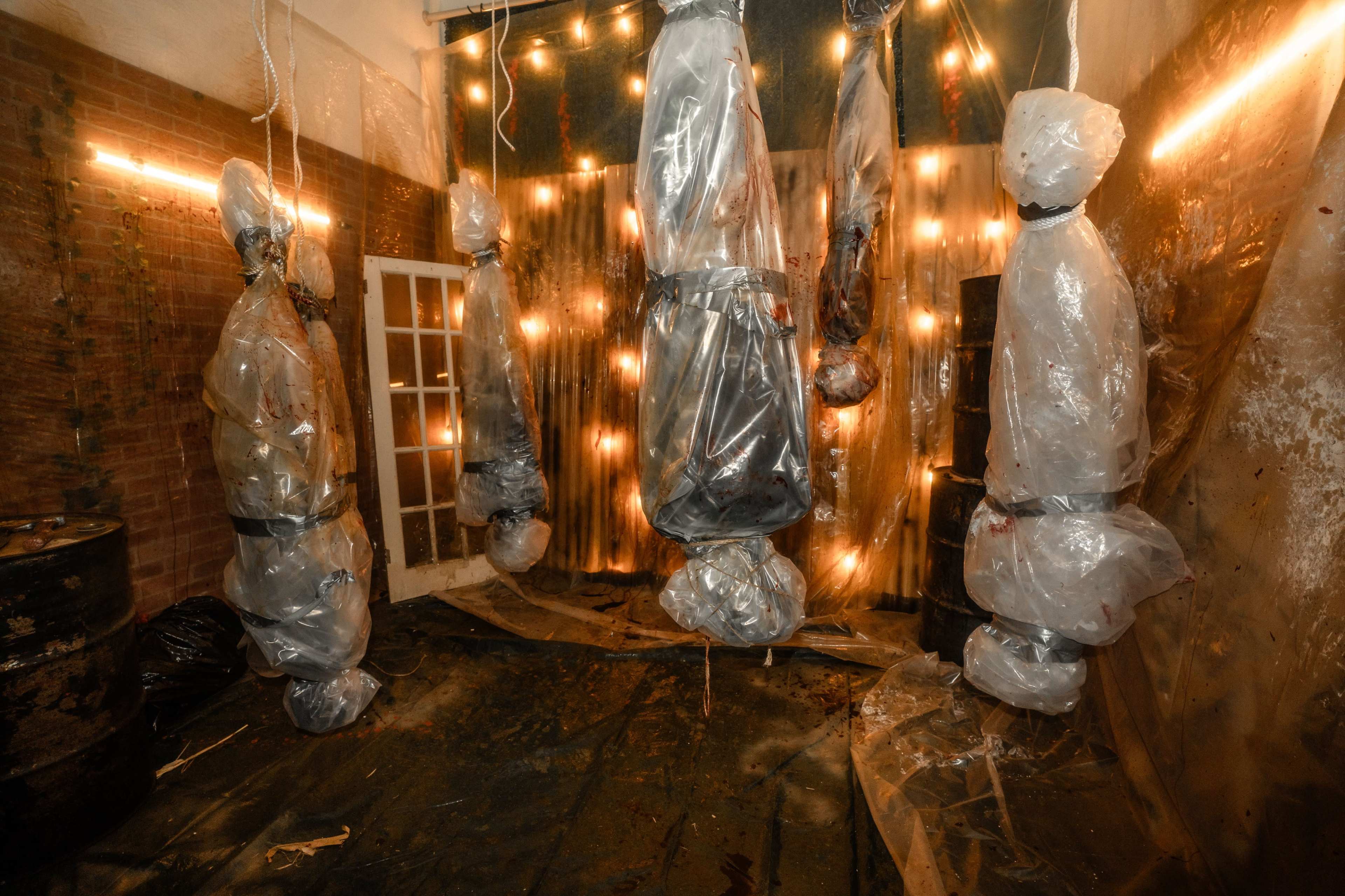 The image depicts a dimly lit room with several figures suspended from the ceiling, each wrapped in clear plastic, surrounded by string lights and a plastic-covered floor.