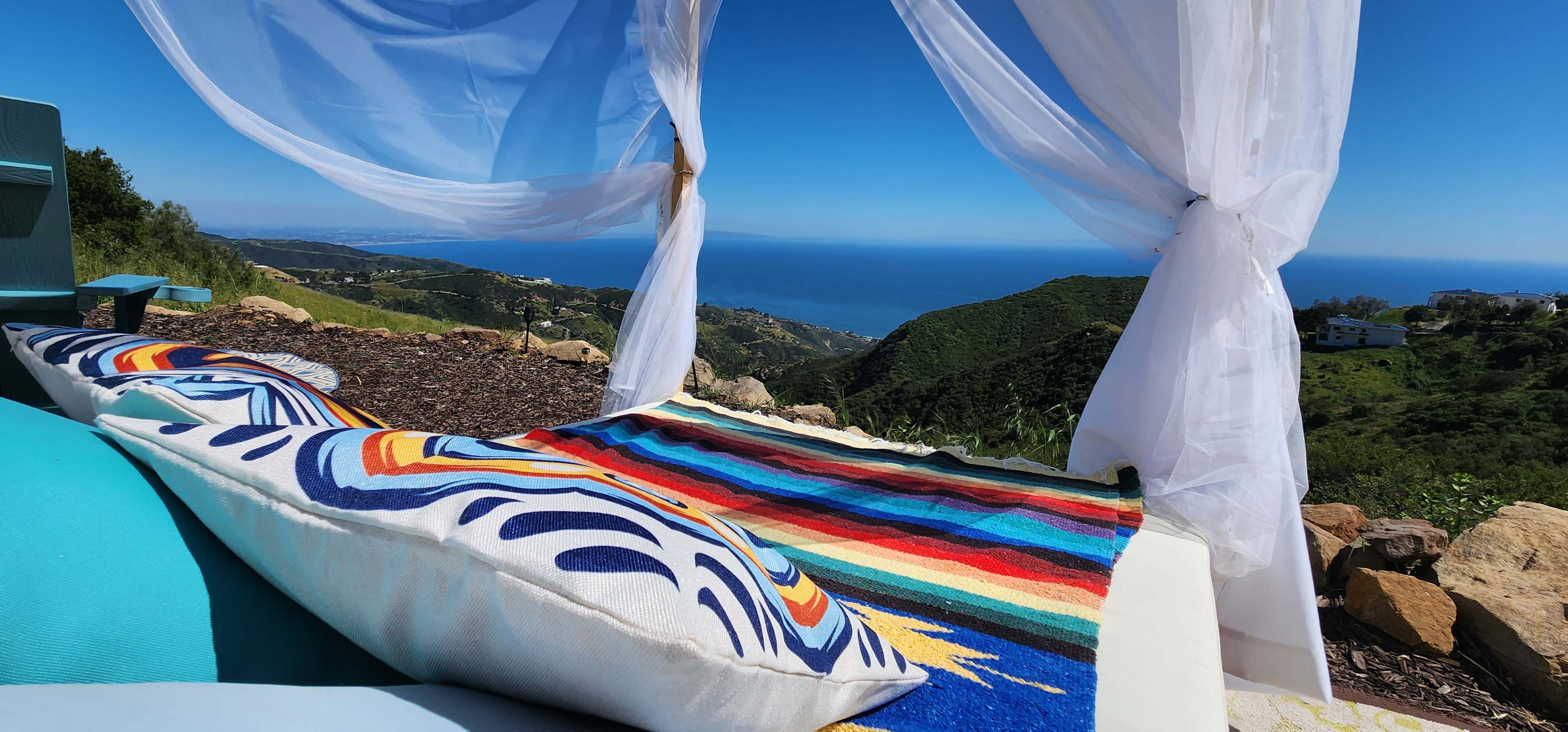 A white canopy drapes over a colorful rug and pillows, overlooking a coastal landscape with the ocean in the distance.
