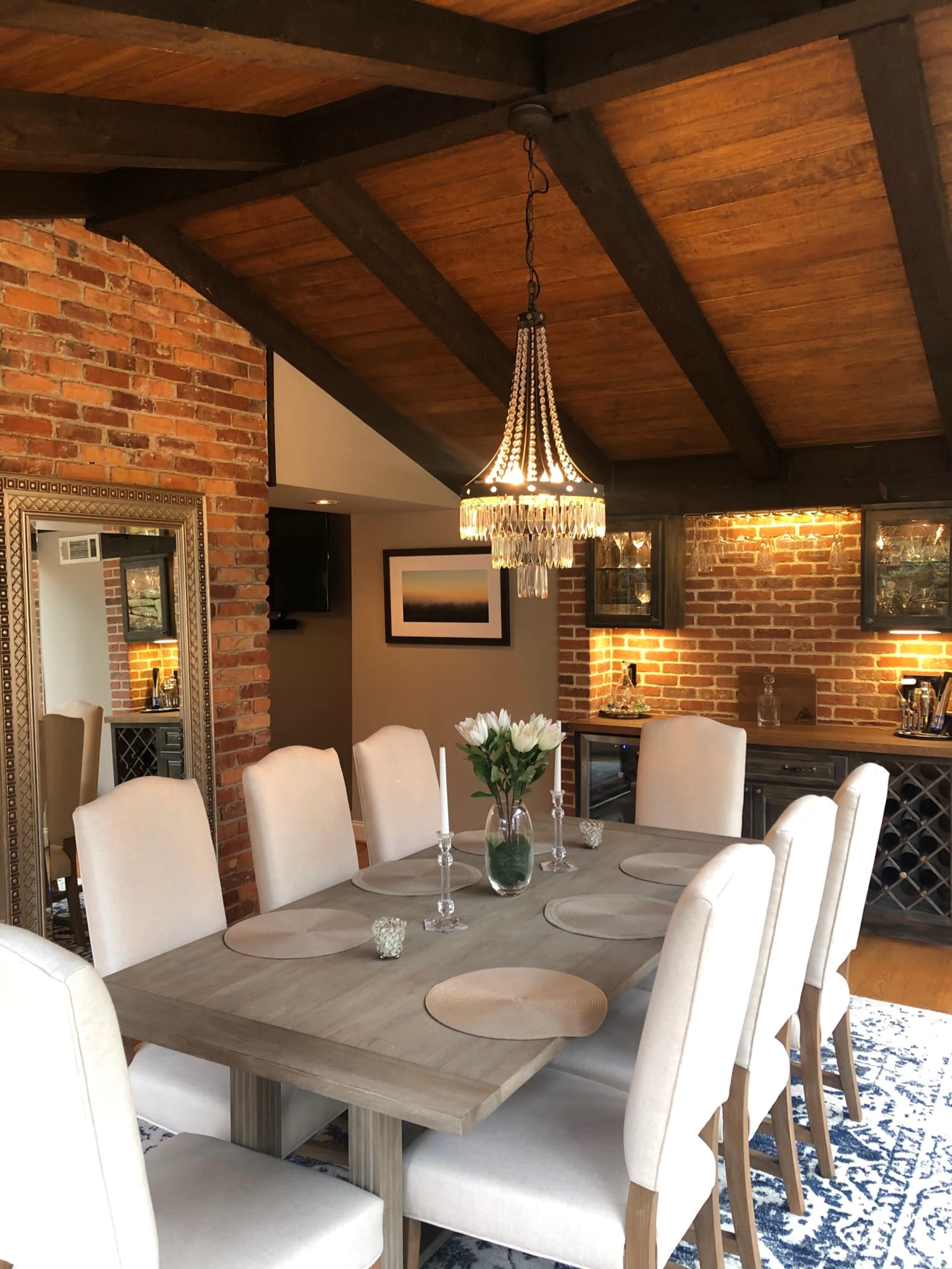 A dining area features a wooden table surrounded by upholstered chairs, with a chandelier overhead and a brick wall in the background.
