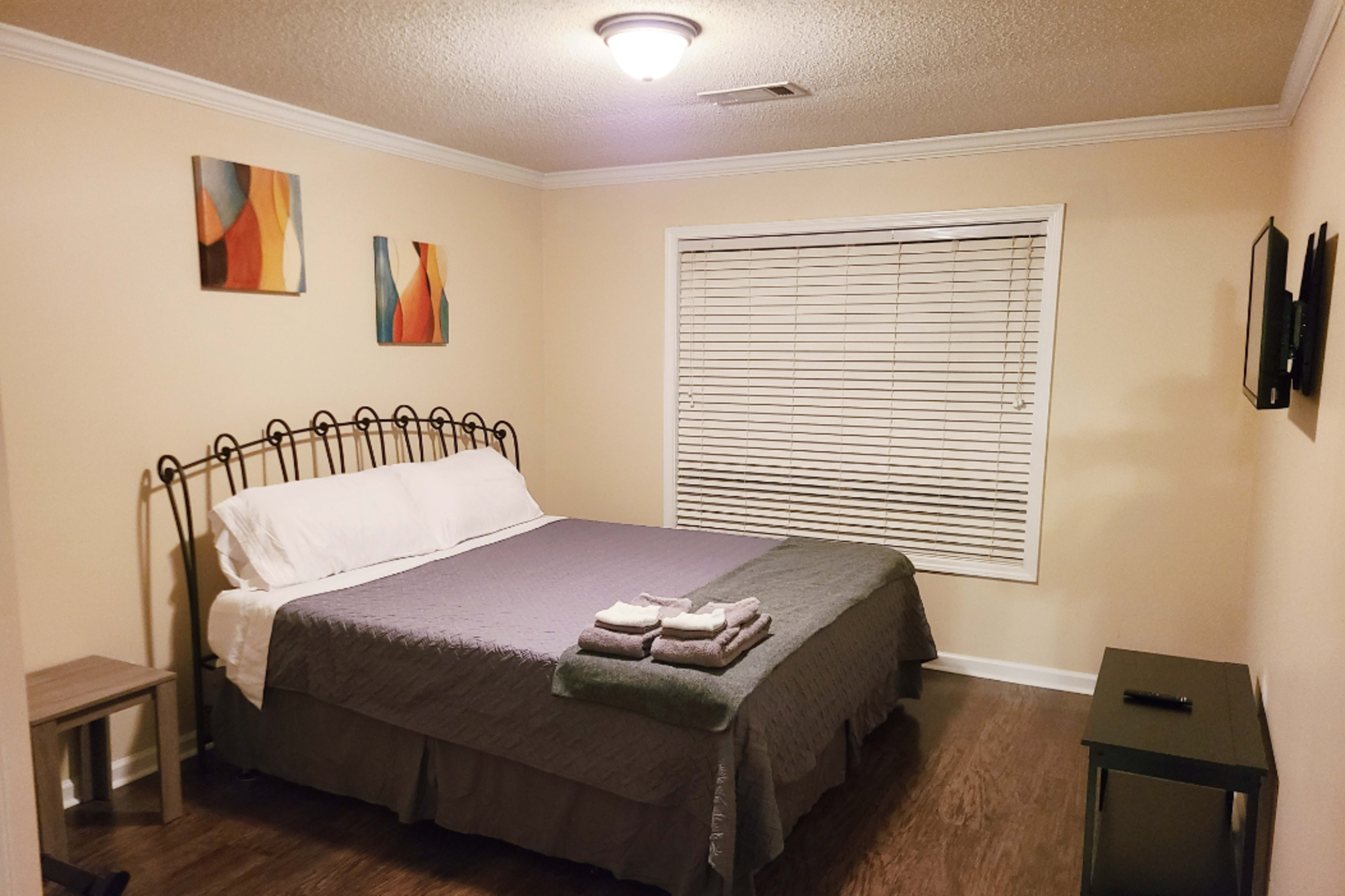 The image shows a cozy bedroom featuring a metal bed frame with a neatly made bed, two pieces of wall art, a small side table, and a television mounted on the wall.