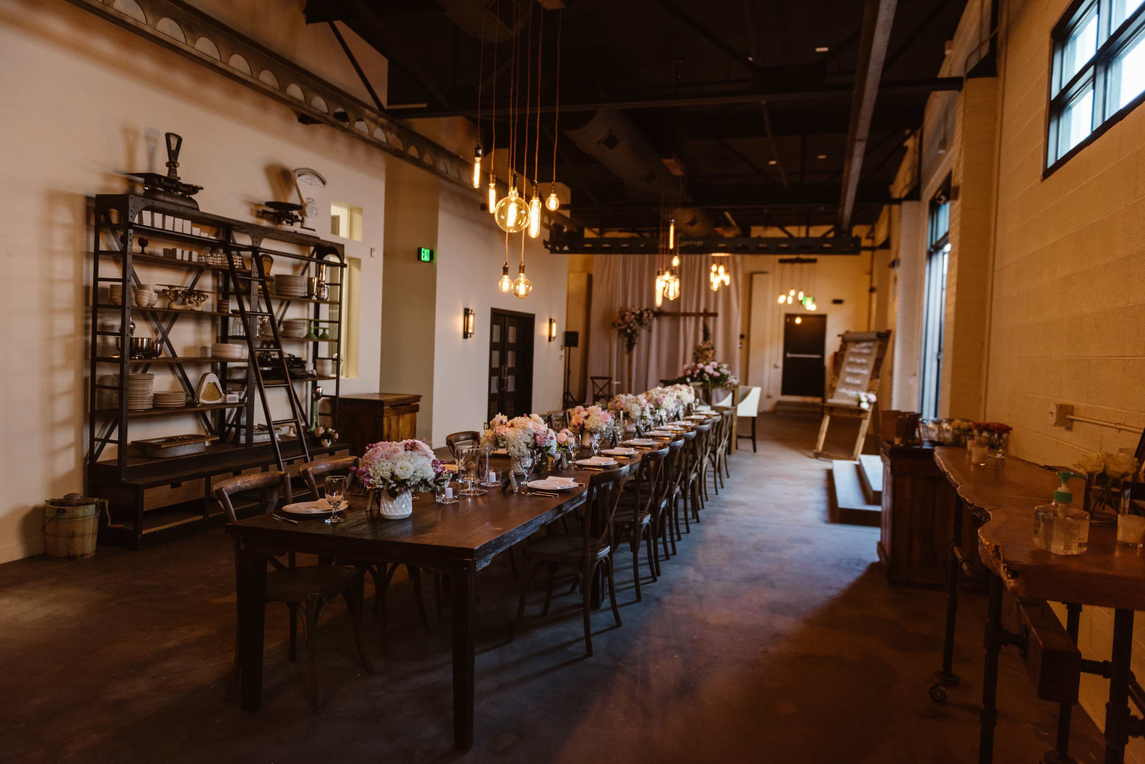 A long wooden dining table is set with plates and floral centerpieces in a spacious, softly lit room featuring industrial decor.