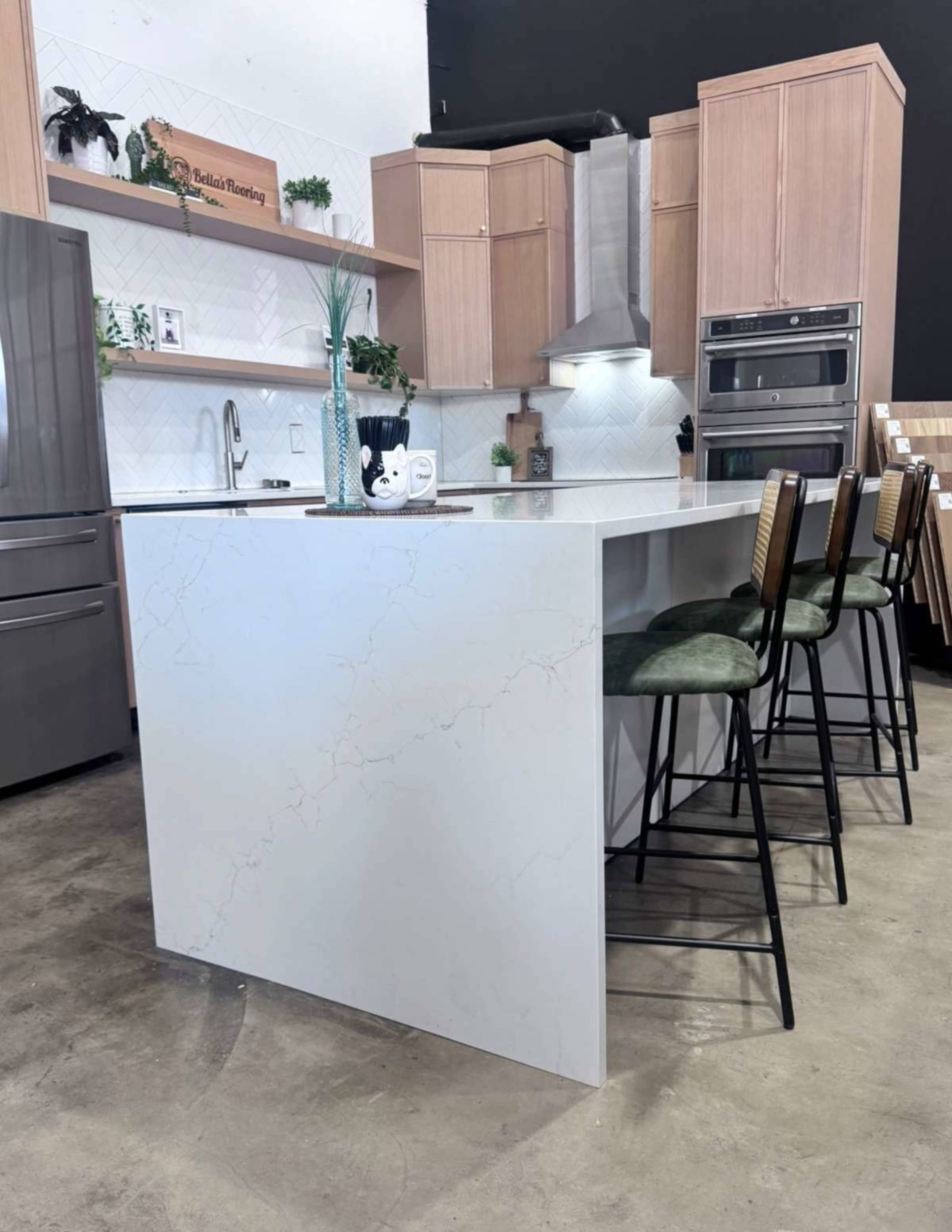The image shows a modern kitchen with a large island featuring a white marble countertop, wooden cabinetry, and four metal bar stools.