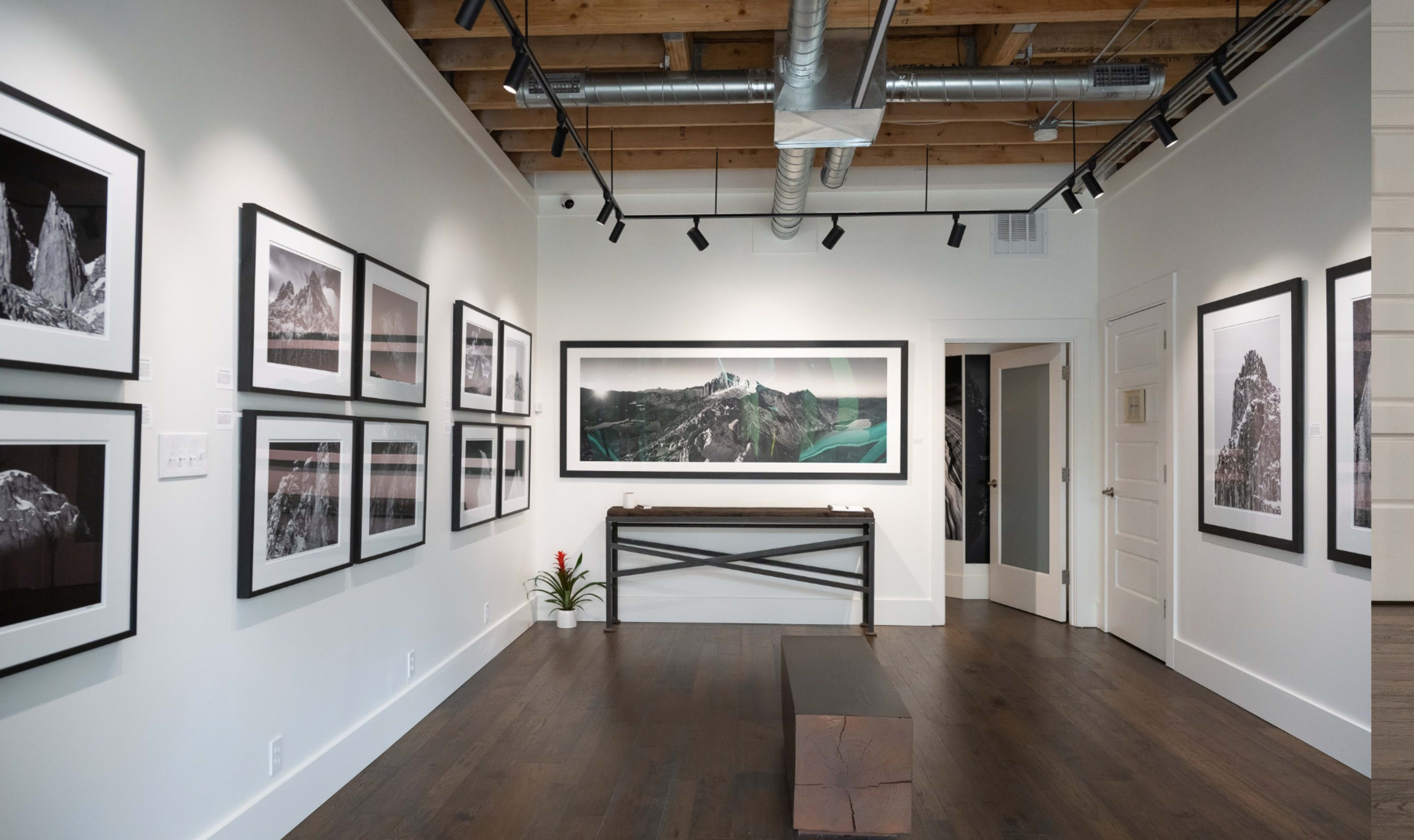 A modern art gallery featuring framed black-and-white photographs on the walls and a large color landscape print at the center, with wooden flooring and a minimalist table.