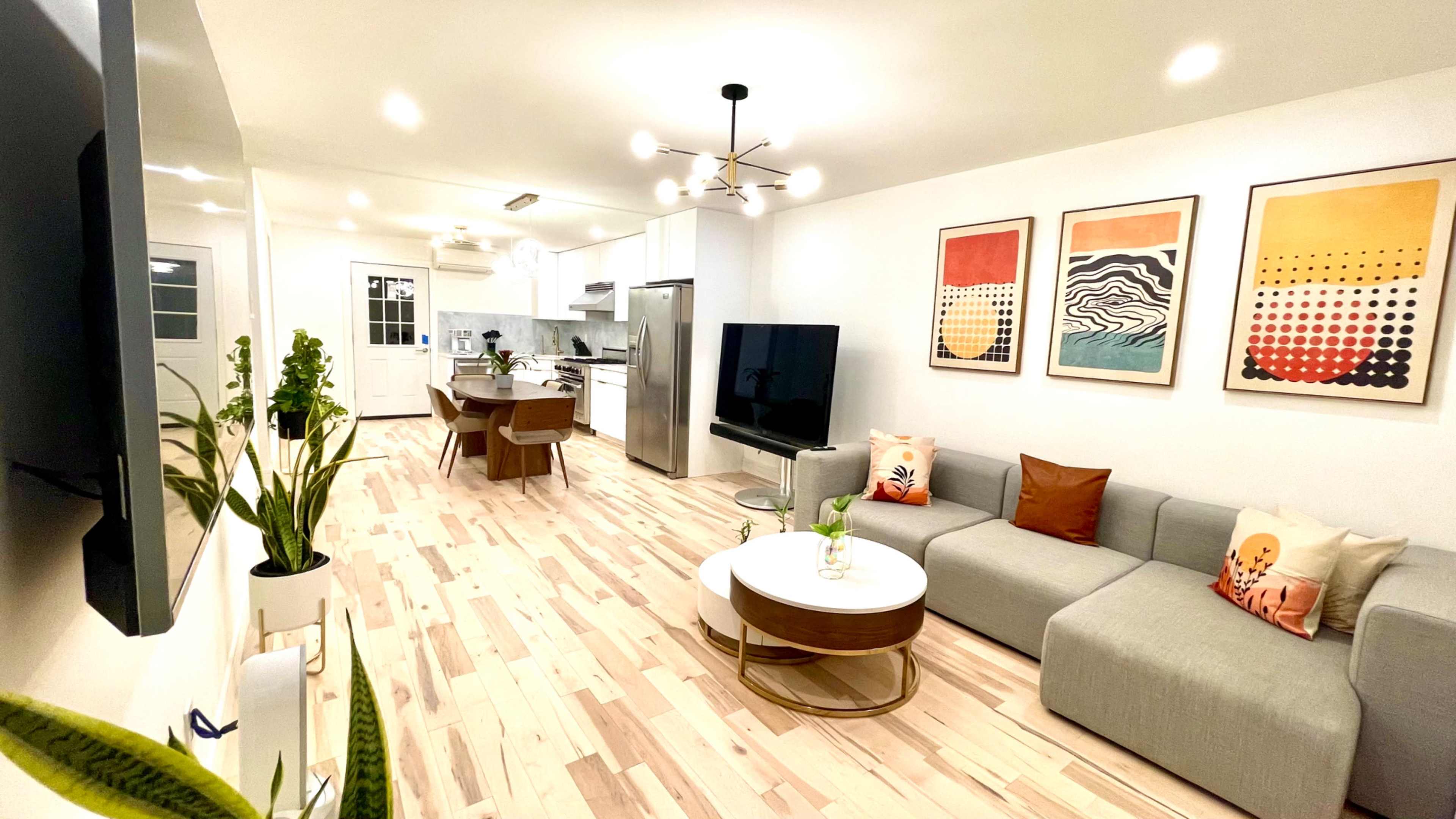 The image shows a modern living room with a gray sectional sofa, a round coffee table, a large TV, and a dining area, all featuring light-colored wood flooring and vibrant artwork on the walls.