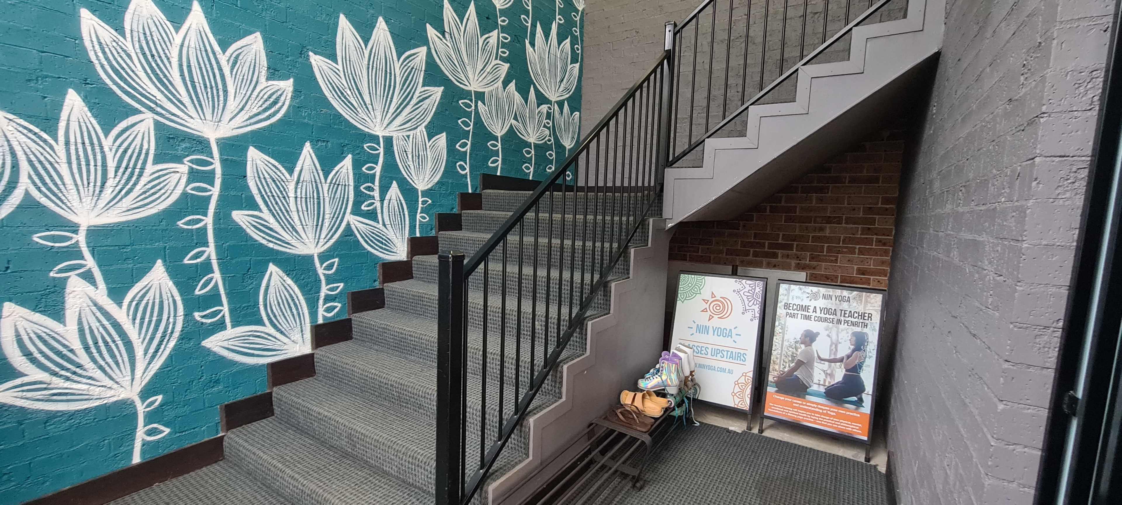 A stairwell with a teal mural featuring white floral designs leads to a landing with informational posters and a bench.