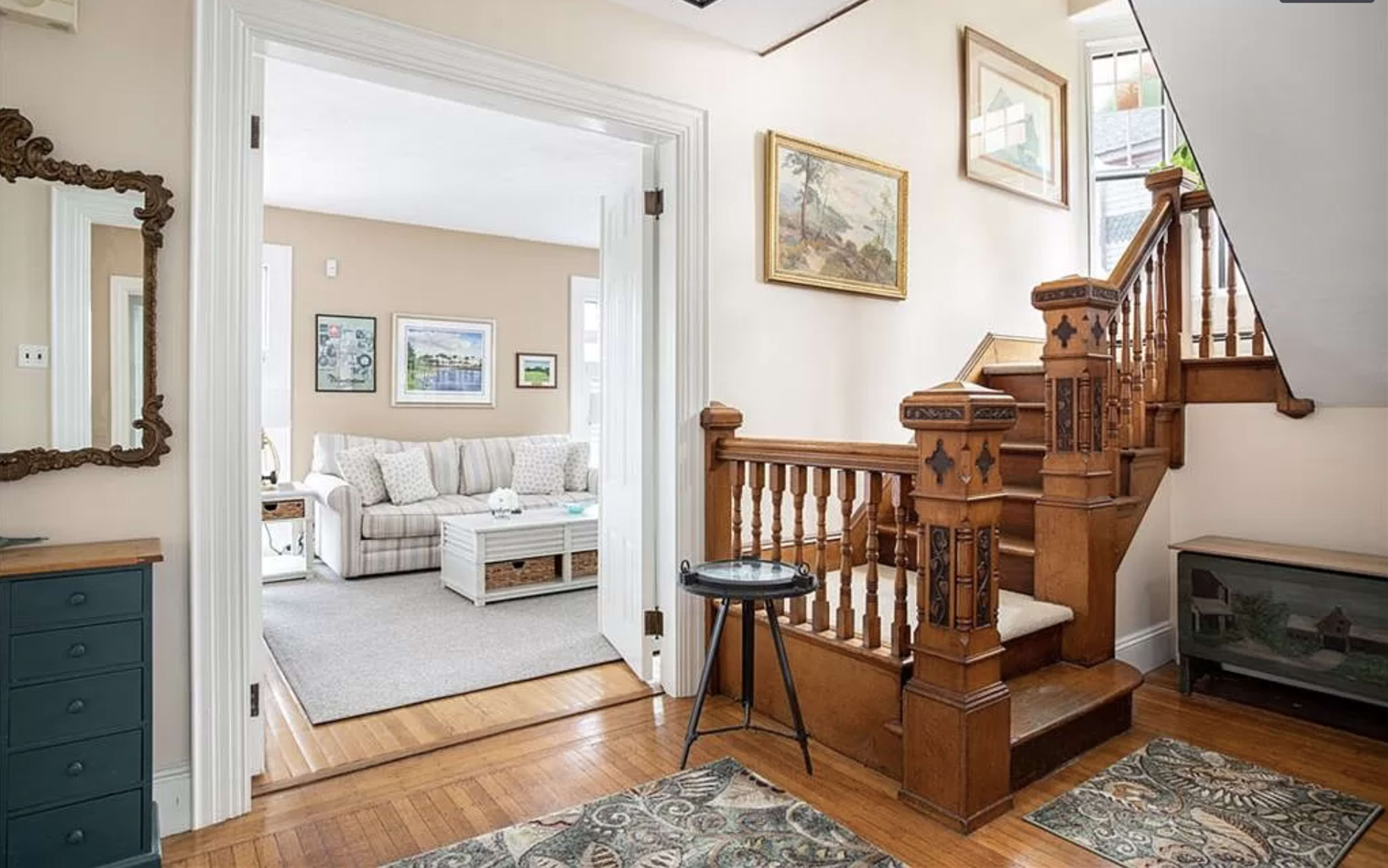 The image shows an entryway featuring a wooden staircase, art on the walls, and a view into a living room with a sofa and coffee table.