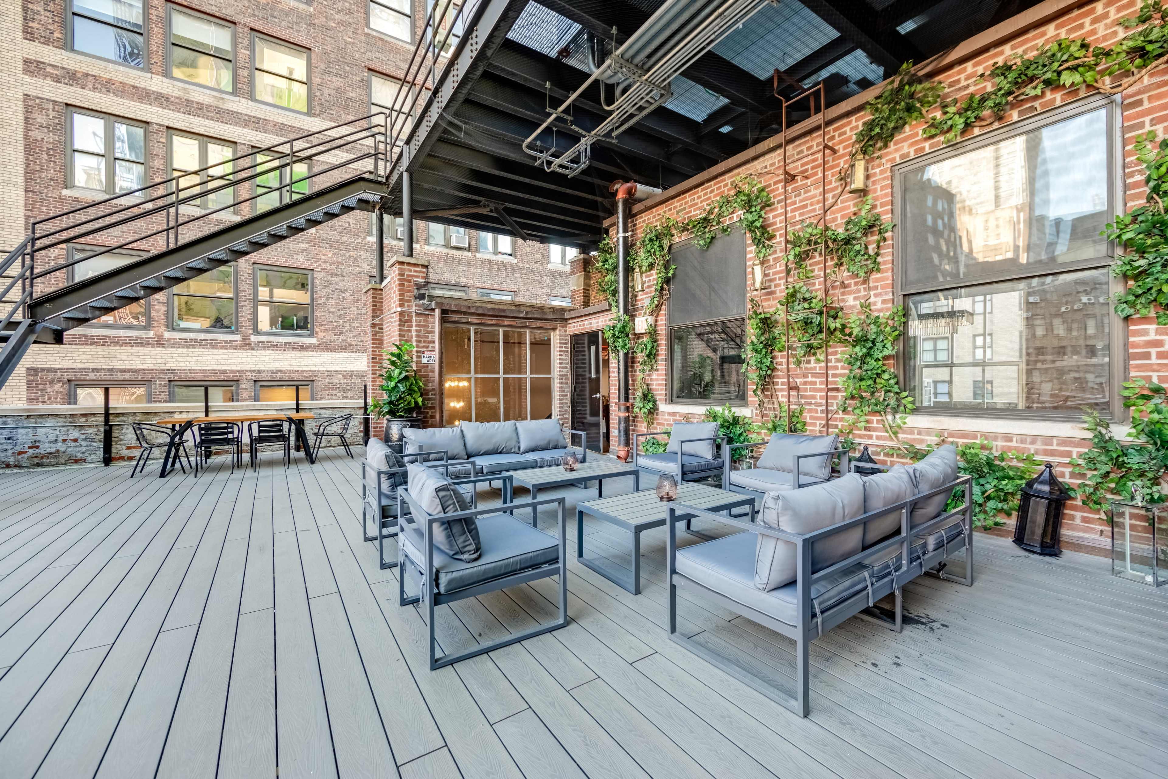 The image shows a rooftop patio with gray seating arrangements, wooden flooring, and brick walls adorned with greenery.