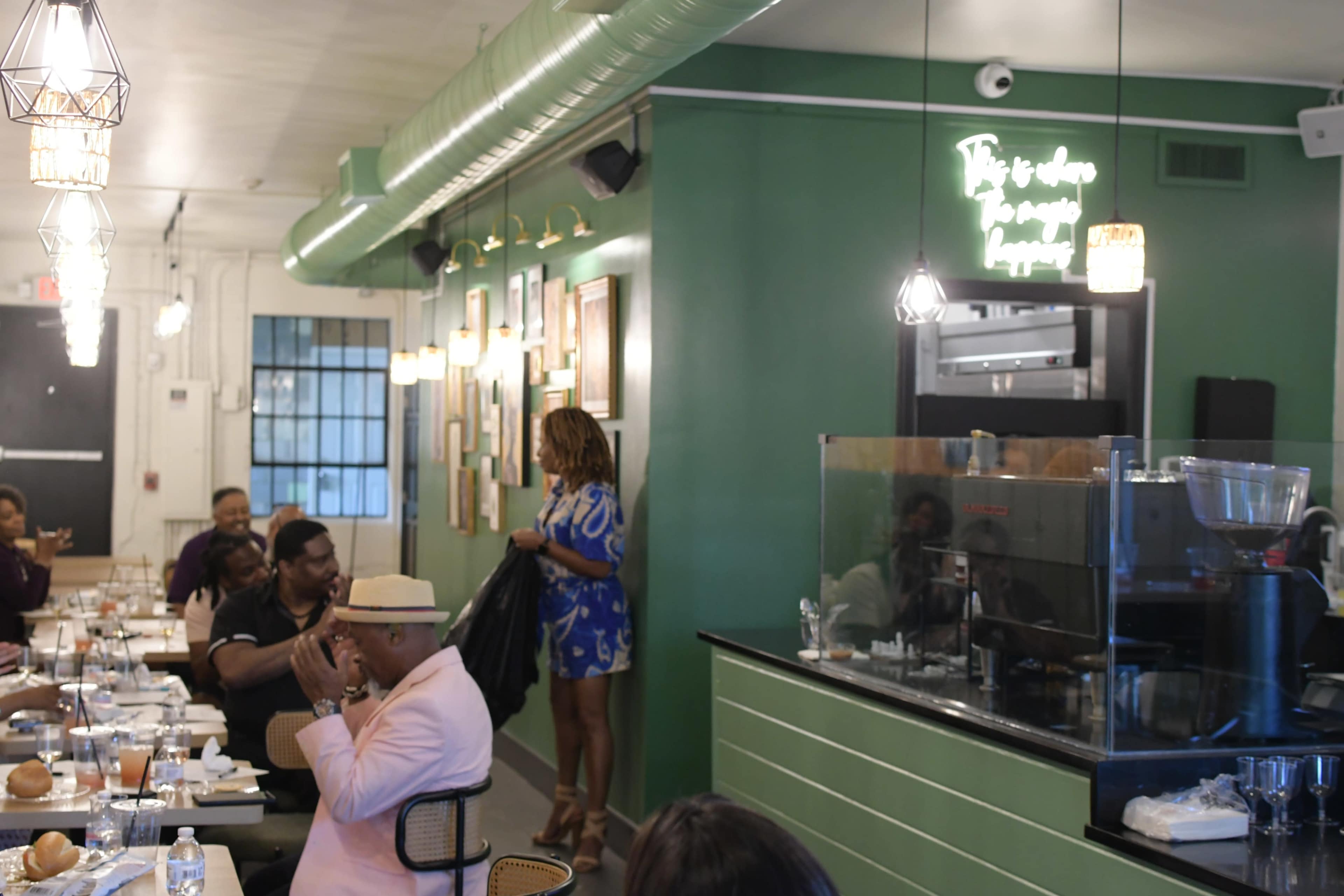 A group of diners sits at tables in a cafe with green walls, while a woman wearing a blue dress stands near the counter.