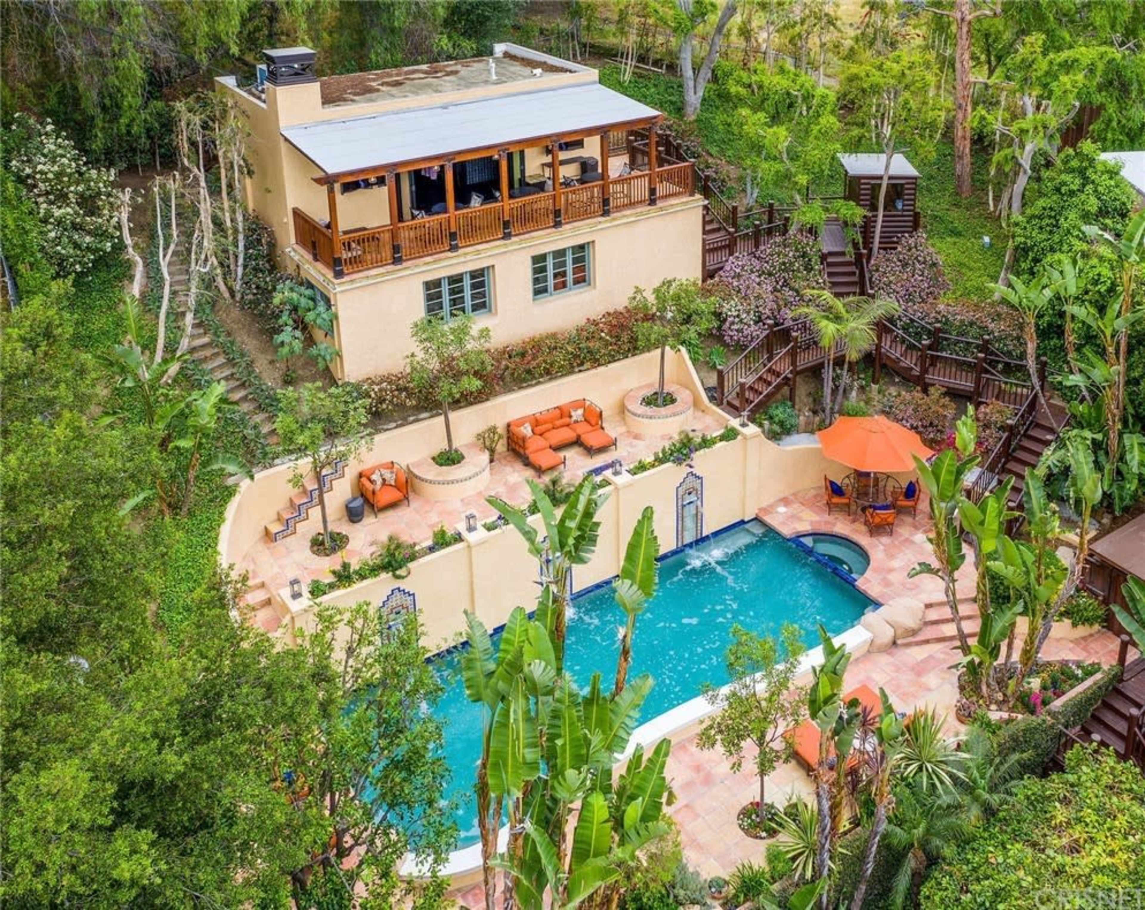 A spacious house with a balcony overlooks a swimming pool surrounded by tropical plants and lounge areas.