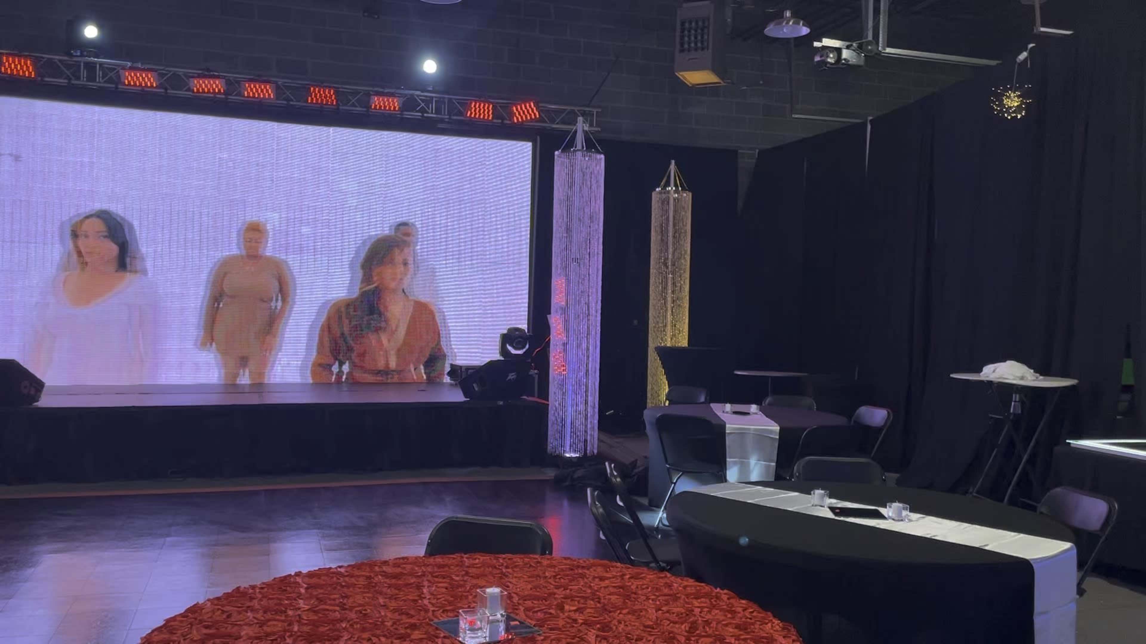 A staged event space featuring a large screen displaying models, decorated tables with red and black tablecloths, and spotlights overhead.