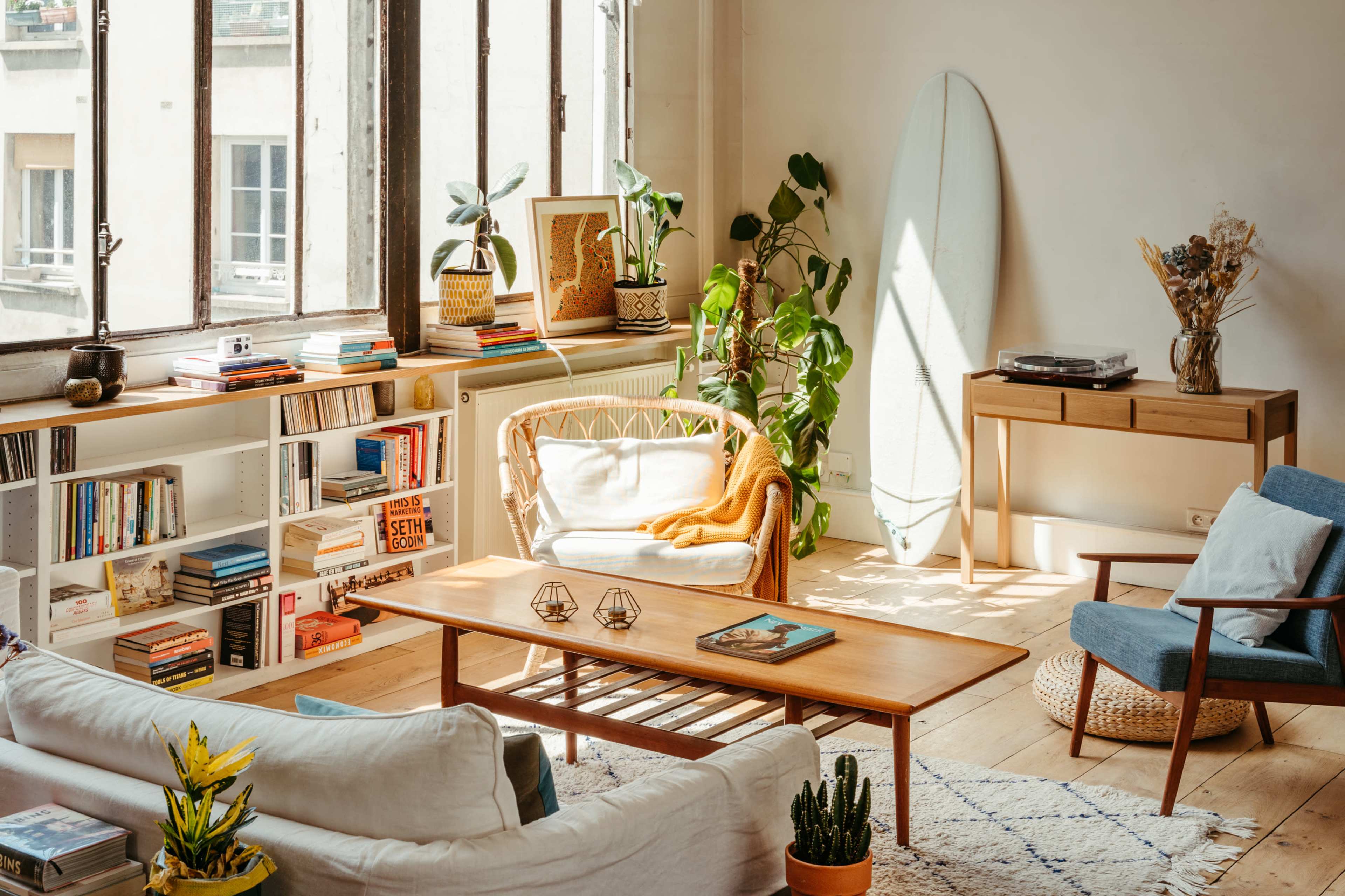 A bright, well-furnished living room with large windows, a coffee table, bookshelves, plants, and a surfboard against the wall.