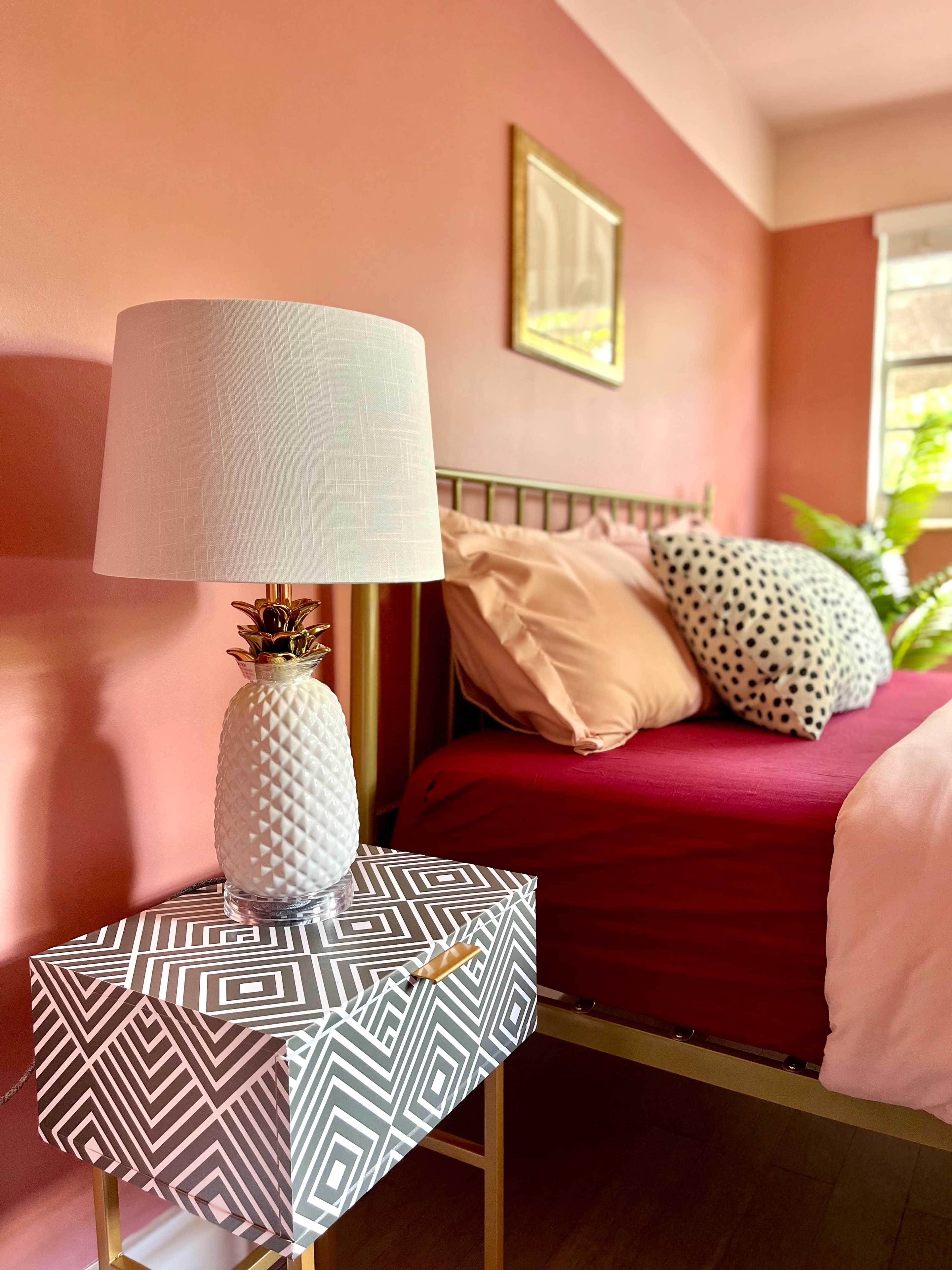 A neatly arranged bedroom features a brass bed frame, a red bedspread, patterned pillows, and a geometric nightstand with a pineapple-shaped lamp.