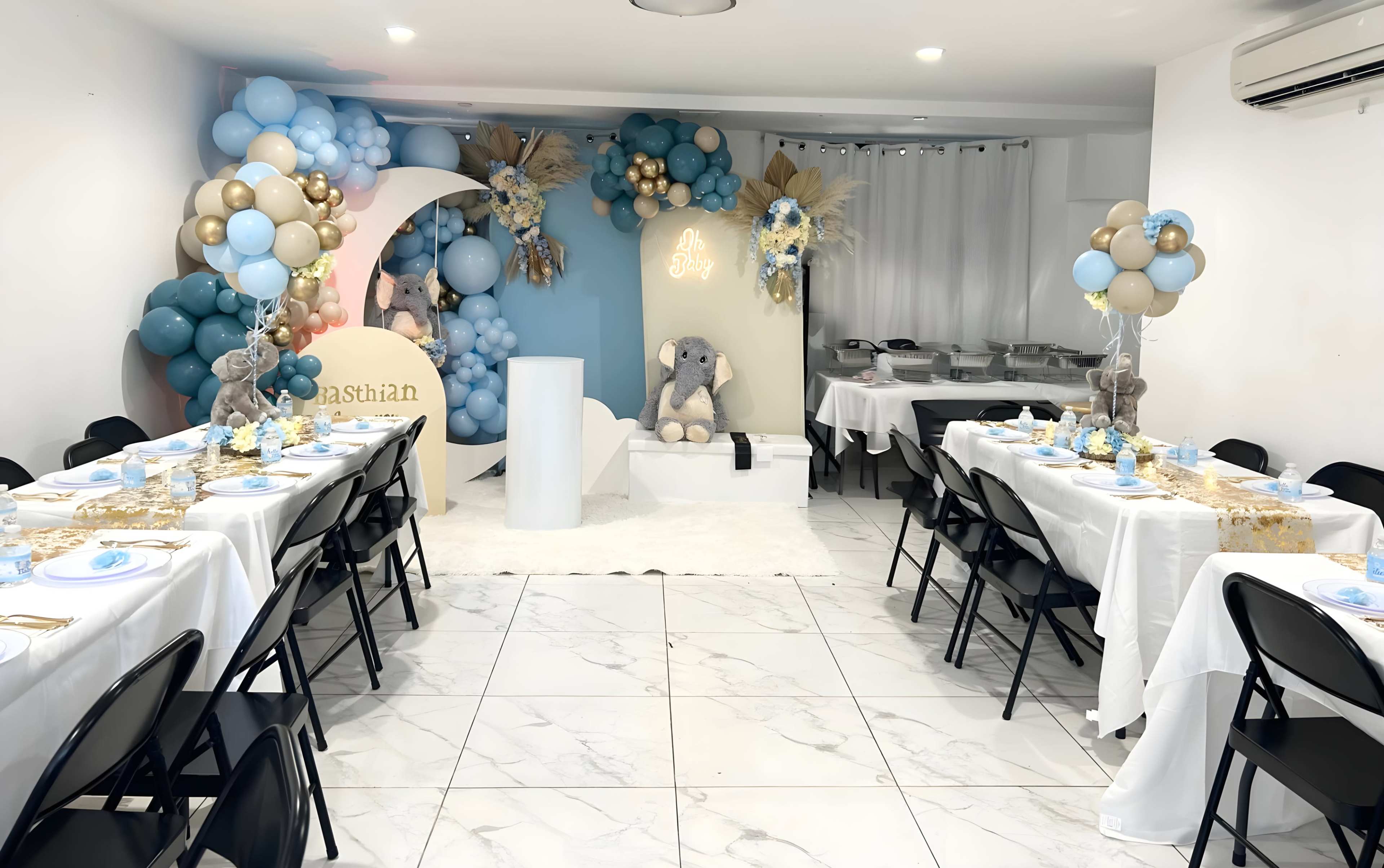 A decorated event space with tables set for a celebration, featuring balloons, a cake display, and a backdrop with a plush elephant theme.
